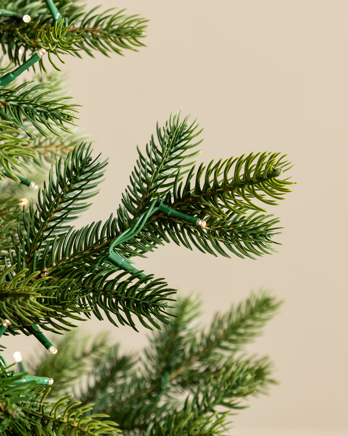 Close-up of the 9' Norway Spruce Pre-Lit Faux Tree, featuring artificial pine branches decorated with small white lights, set against a plain beige background, by Vendixs