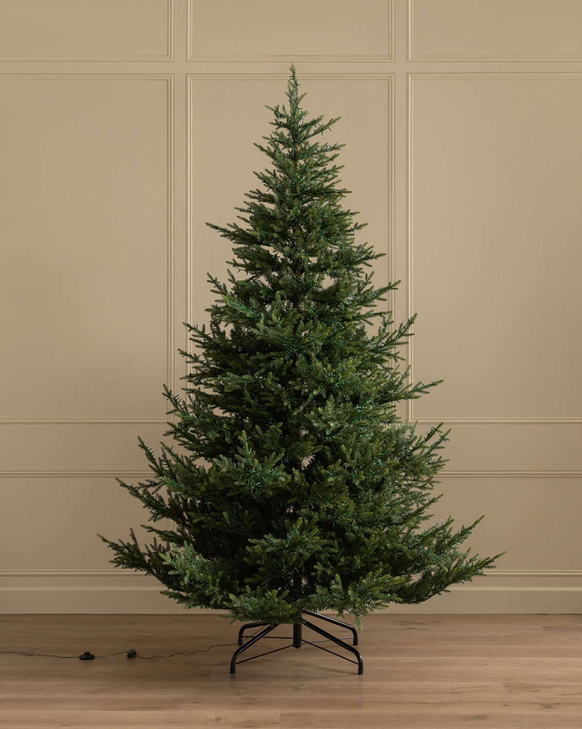 A 9' Norway Spruce Pre-Lit Faux Tree with dense green branches stands on a metal stand in a plain room with beige paneled walls and wooden floor, an electric cord lying nearby, by Vendixs
