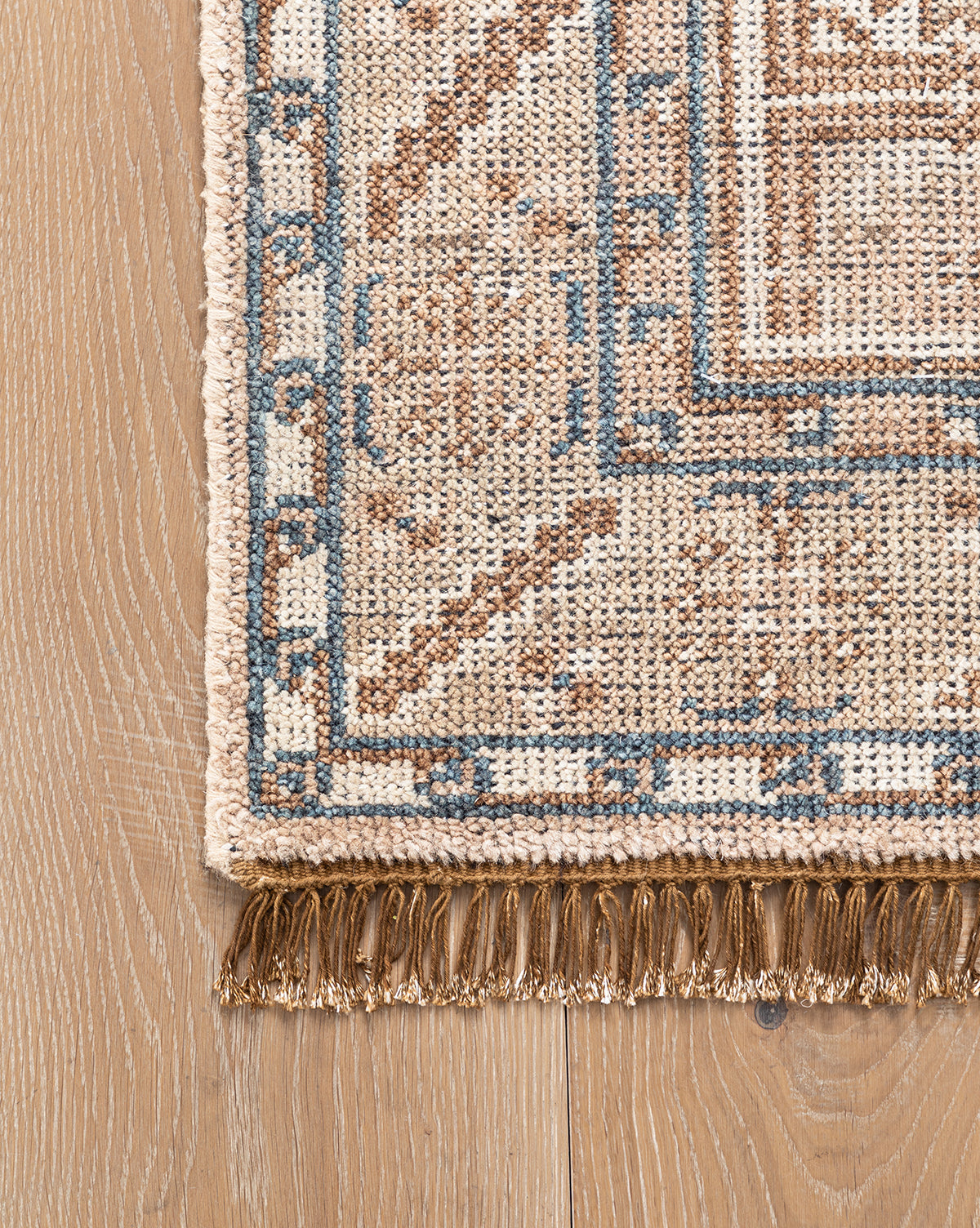 Close-up of the Nunez Hand-Knotted Wool Rug shows its beige, brown, and blue geometric pattern, tasseled fringe, and classic border design, displayed on a light wooden floor, by Vendixs