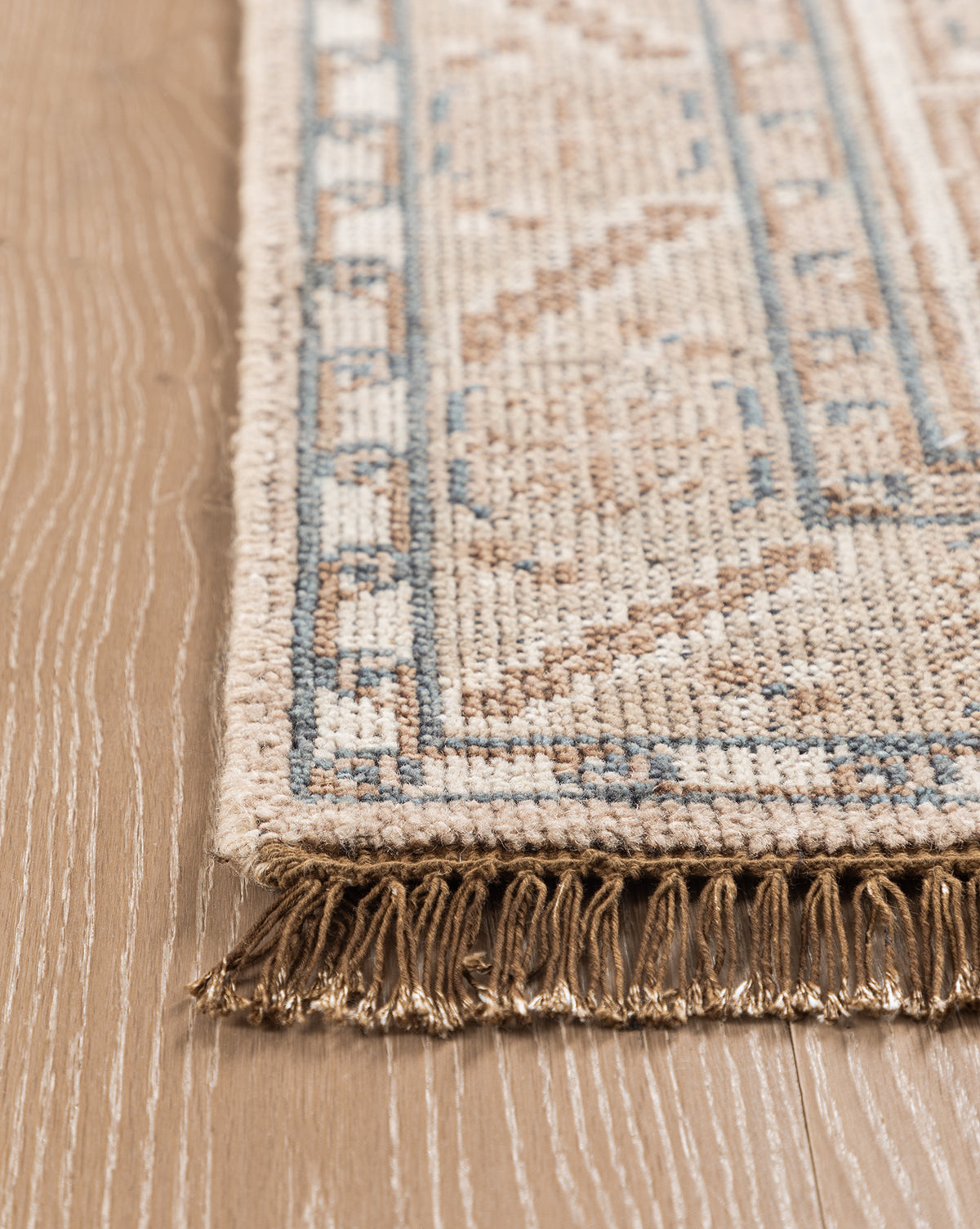 A close-up of the Nunez Hand-Knotted Wool Rug shows its beige and blue geometric pattern, fringed edges, and classic border design, beautifully placed on a light wood floor, by Vendixs