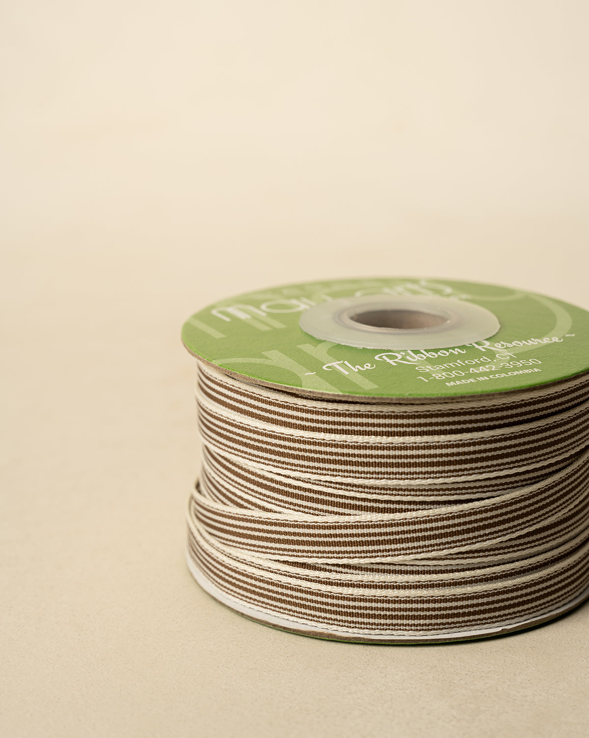 A spool of 3/8" Oatmeal & Brown Woven Grosgrain Stripe Ribbon, neatly wound with a green label on top, rests on a beige surface—ideal for holiday decor or gift wrapping, by Vendixs
