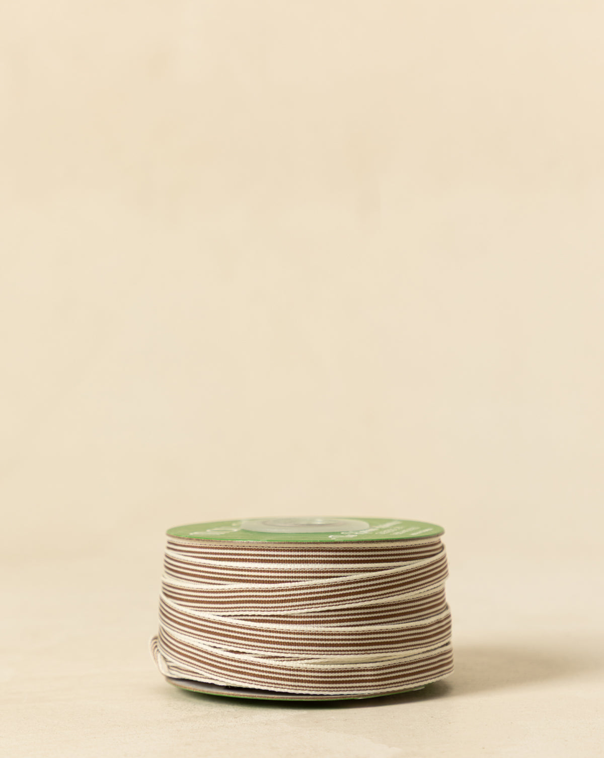 A spool of 3/8" Oatmeal & Brown Woven Grosgrain Stripe Ribbon rests on a light beige surface with a plain background—ideal for holiday decorating, by Vendixs