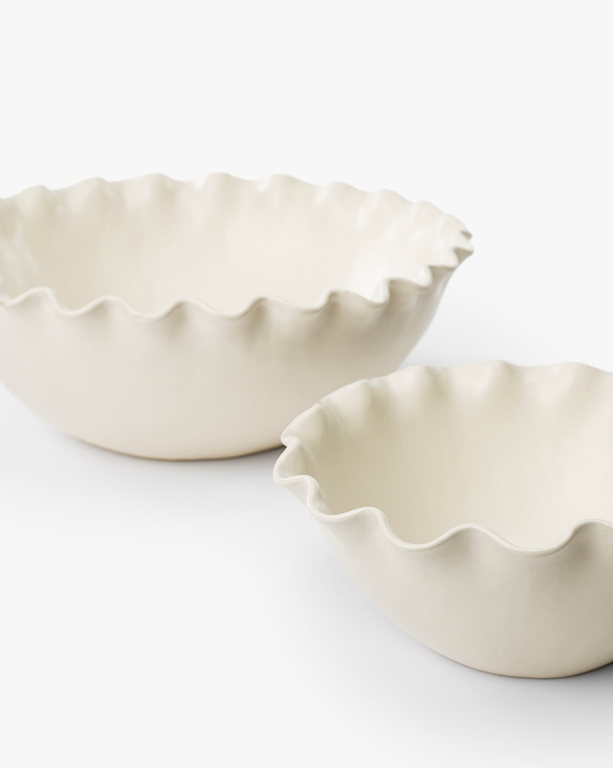 Two off-white Olindo Serving Bowls (Set of 2) with ruffled, wavy edges are placed side by side on a plain white surface, one slightly behind the other. These vintage-inspired bowls are empty by Vendixs