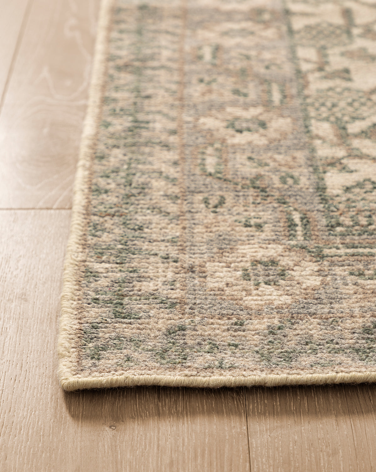 Close-up of the Orsino Hand-Knotted Rug, featuring green and beige patterns, lying flat on a light wood floor. Its antique-style design is highlighted by a neatly stitched border along the corner, by Vendixs