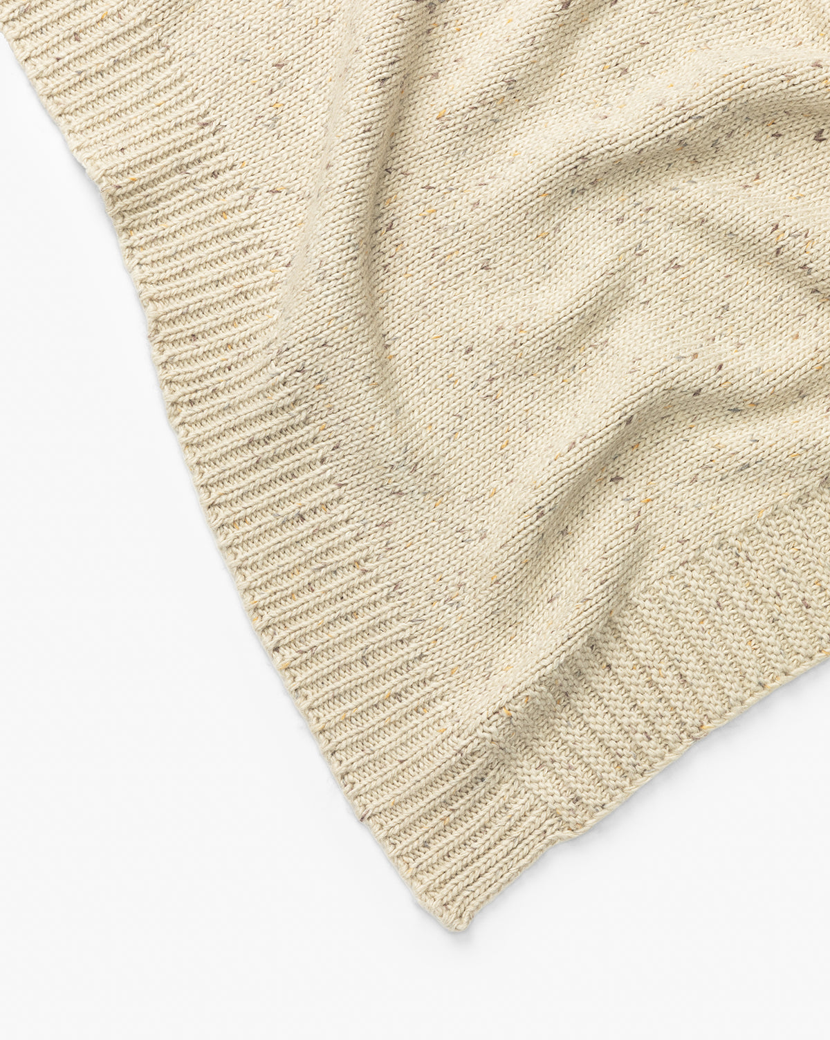 A close-up of the Makerspalm Otis Knit Throw shows its beige wool blend with ribbed edges, a subtly wavy texture, and small grey-speckled flecks throughout. Vendixs