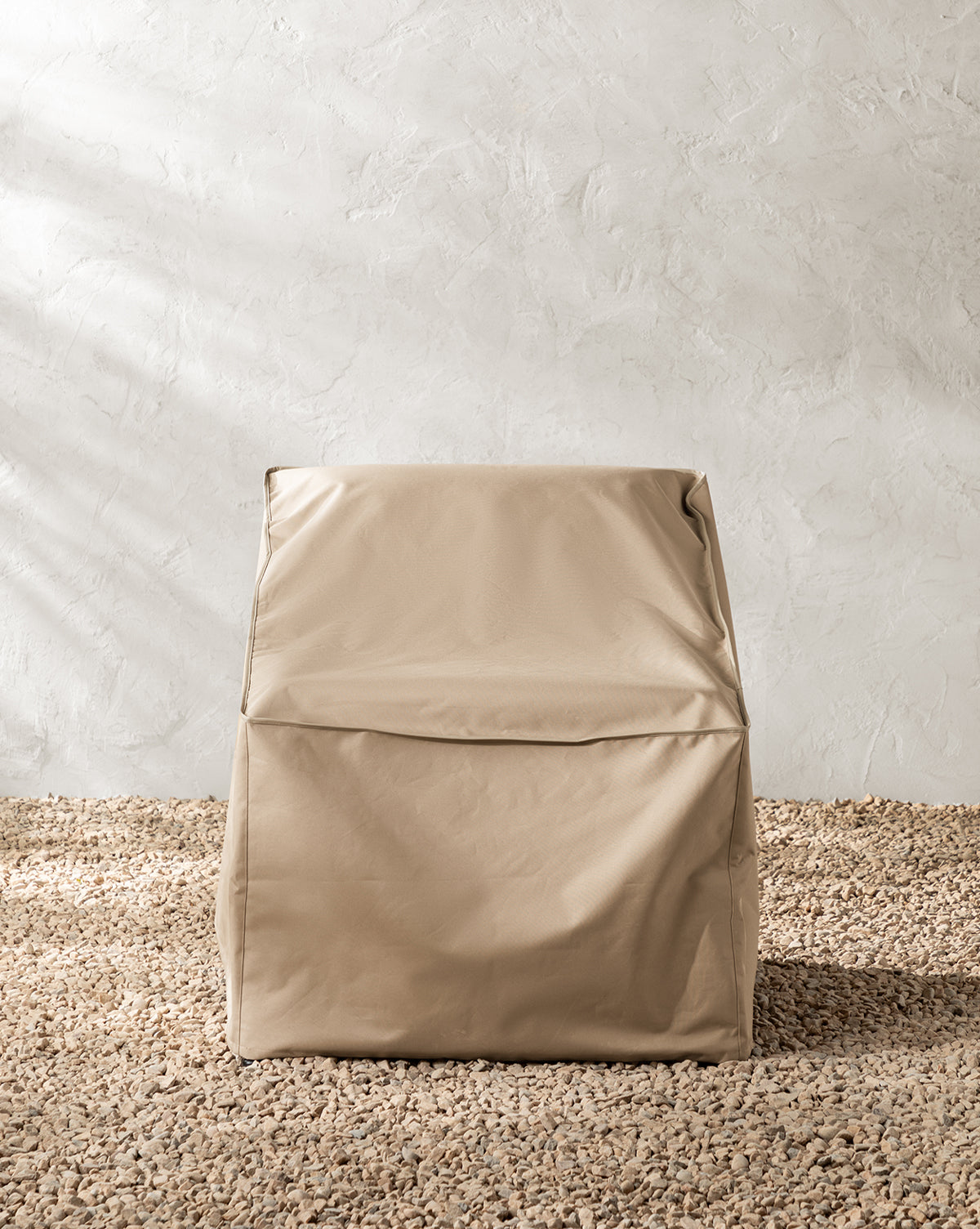A beige, boxy chair with a leather-like texture sits on pebbled flooring against a light wall, shielded from the elements by the water-resistant Outdoor Lounge Chair Cover, by Vendixs