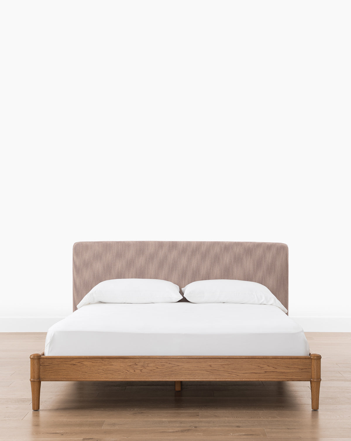 The Palladio Bed by Makerspalm features a wooden frame with honey oak legs and a light brown upholstered headboard, dressed in white sheets and pillows, set on a wooden floor against a plain white wall, finished with accents from Vendixs.