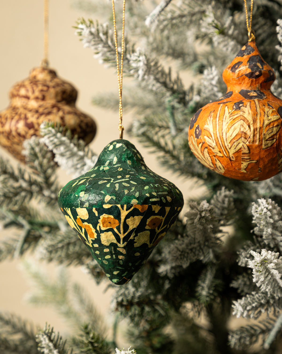Eight hand-painted, block-printed paper mâché ornaments from the Paper Mache Block Printed Ornament Set add floral patterns in green, orange, and brown to a snow-dusted Christmas tree branch, by McGee & Co.