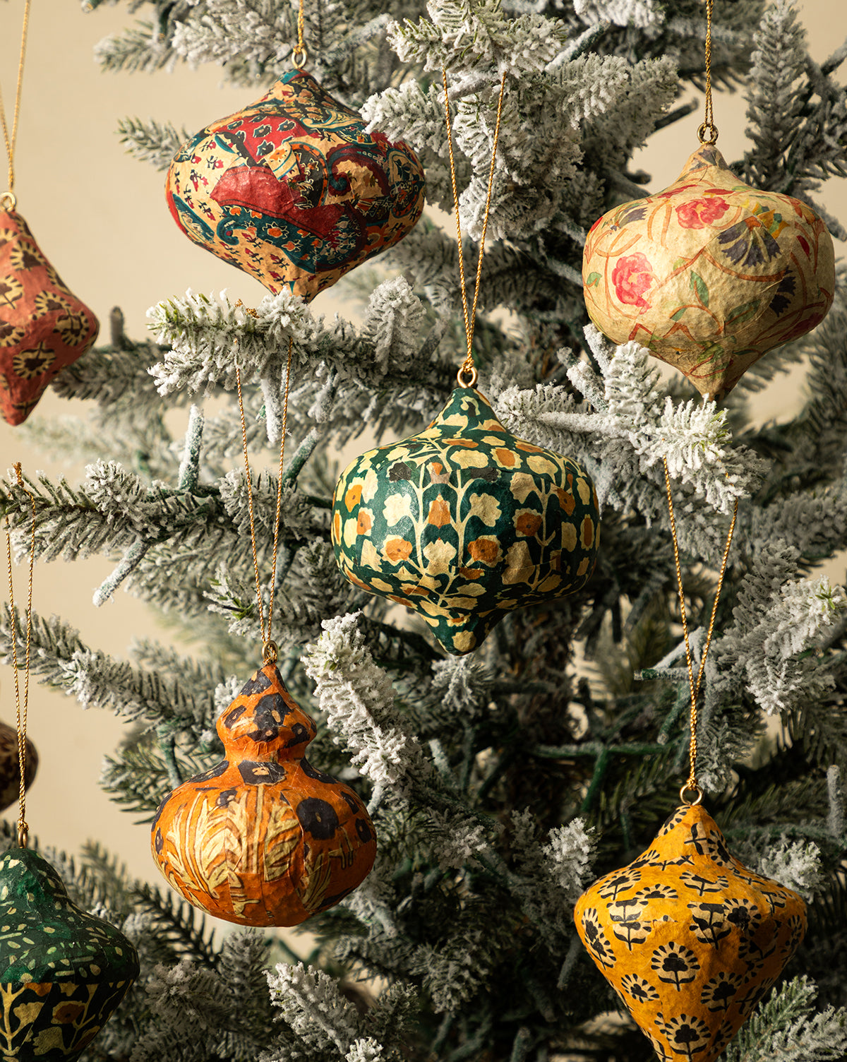 The Paper Mache Block Printed Ornaments (Set of 8) hang from a snow-frosted Christmas tree, displaying vibrant green, yellow, red, and gold patterns, by Vendixs