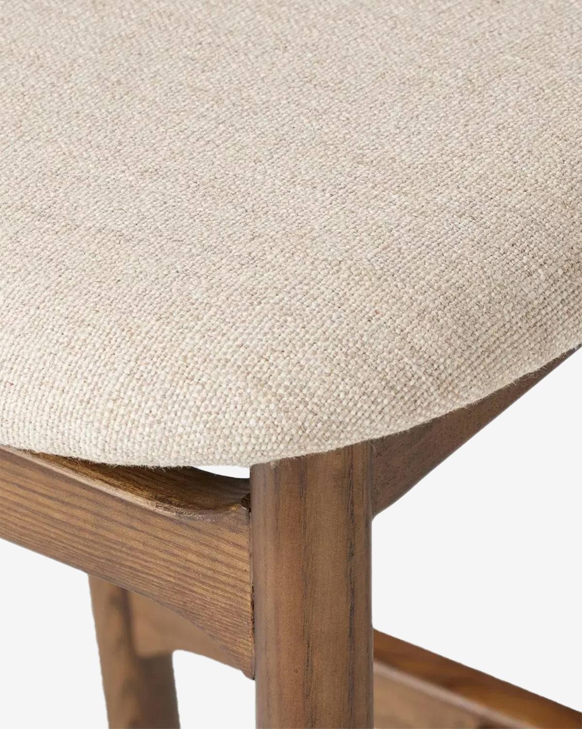Close-up of the Patrin Stool by Four Hands, showcasing its beige textured performance upholstery and sturdy ash wood frame in a natural brown finish, with details of the seat and leg joint visible. Vendixs
