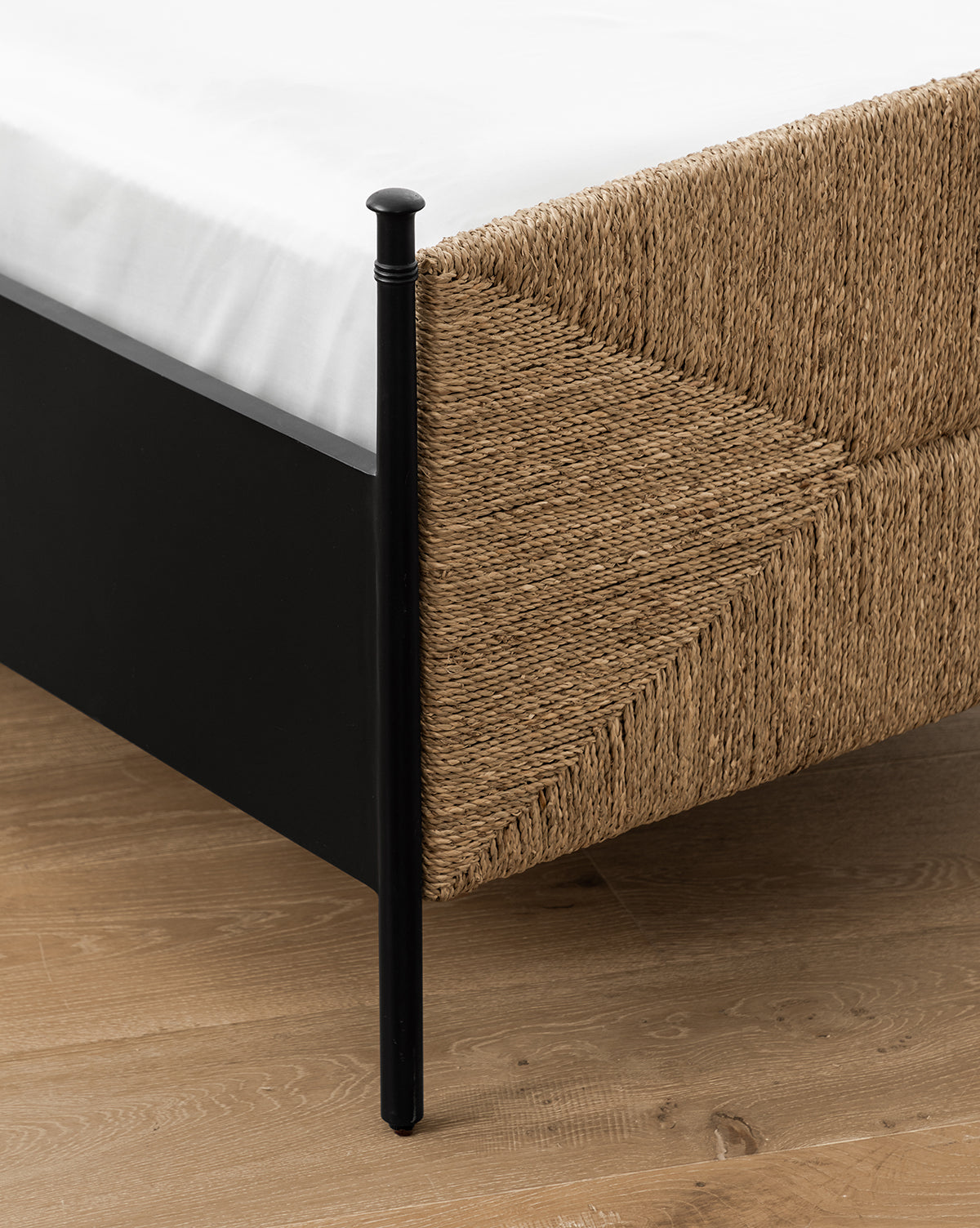 Close-up of the Peregrine Bed’s black metal frame and woven seagrass headboard at the corner, shown on a light wood floor with a white mattress on top, by Vendixs