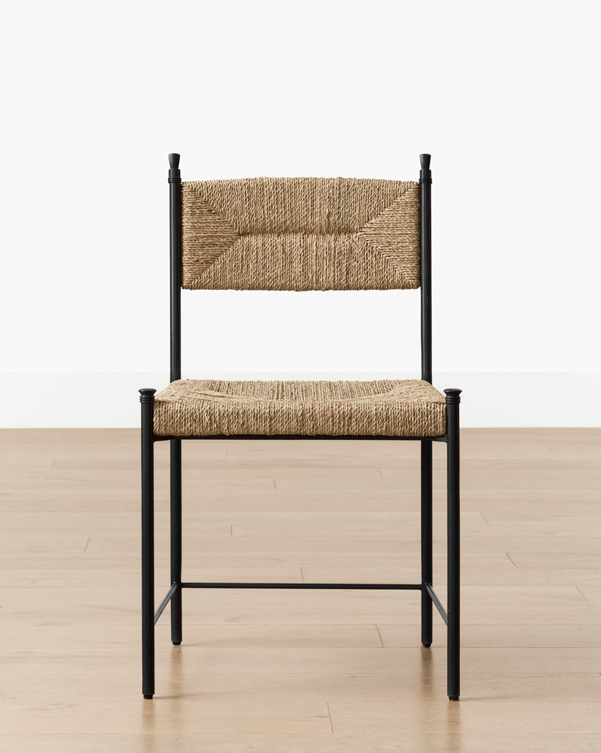 A Peregrine Chair featuring a black metal frame and woven natural fiber seat and backrest sits on a light wood floor before a plain white wall, by Vendixs