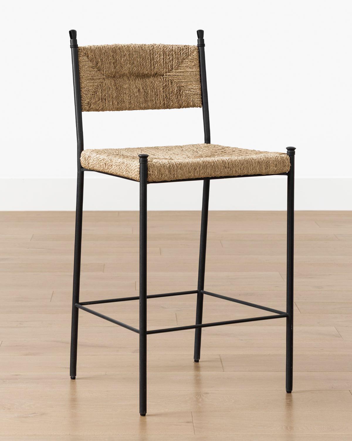 The Peregrine Stool by Makerspalm features a black steel frame with woven natural rattan seat and backrest, set on a light wooden floor against a plain white wall. Vendixs