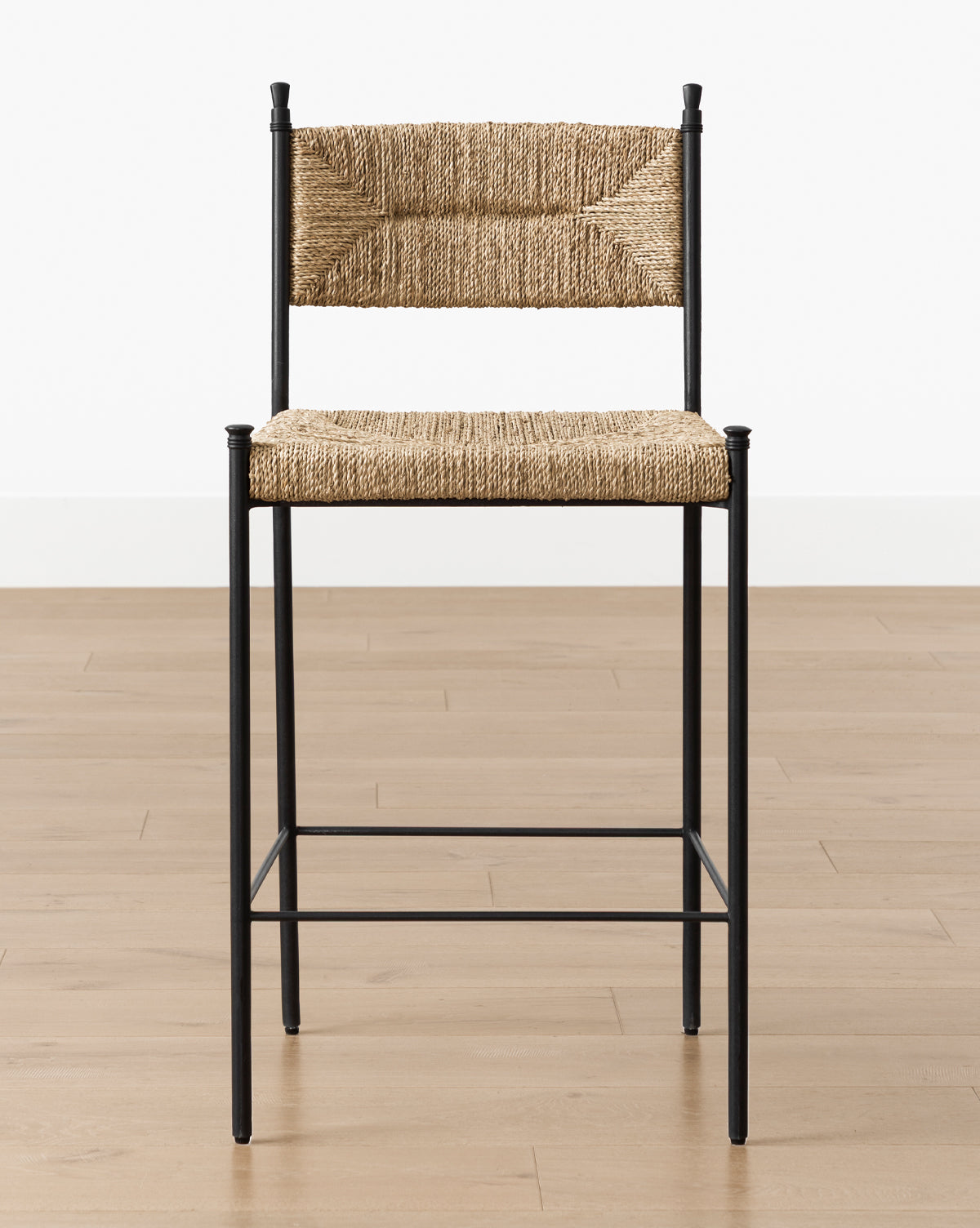 The Makerspalm Peregrine Stool, featuring a black metal frame with woven seagrass seat and backrest, sits on a light wood floor against a white wall—perfect as a stylish kitchen dining stool. Vendixs