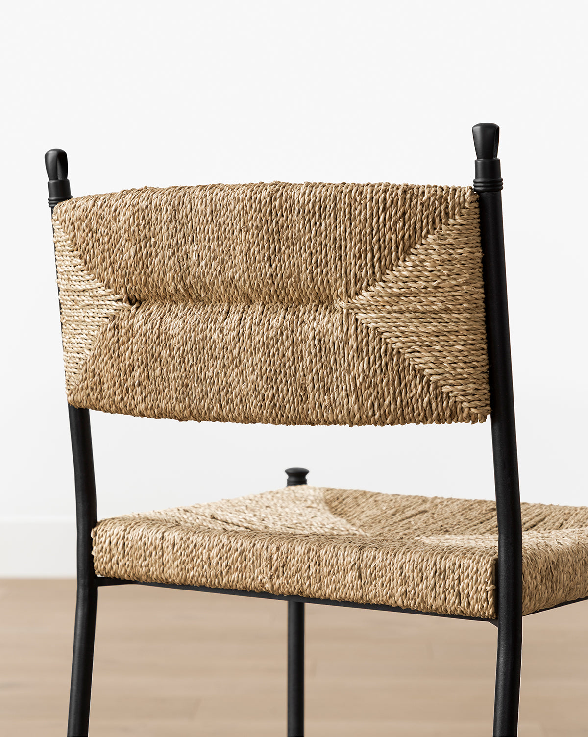 The Peregrine Stool by Makerspalm features a black steel frame with woven seagrass seat and backrest in natural fibers, shown against a plain white wall and wooden floor. Vendixs