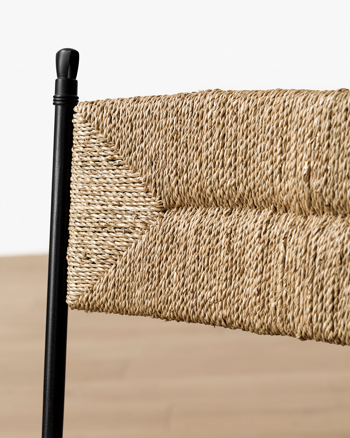 Close-up of the Peregrine Stool by Makerspalm—featuring a chevron-patterned woven seagrass and natural rope backrest, black steel frame, and blurred light wood floor background—ideal as a stylish kitchen dining stool. Vendixs