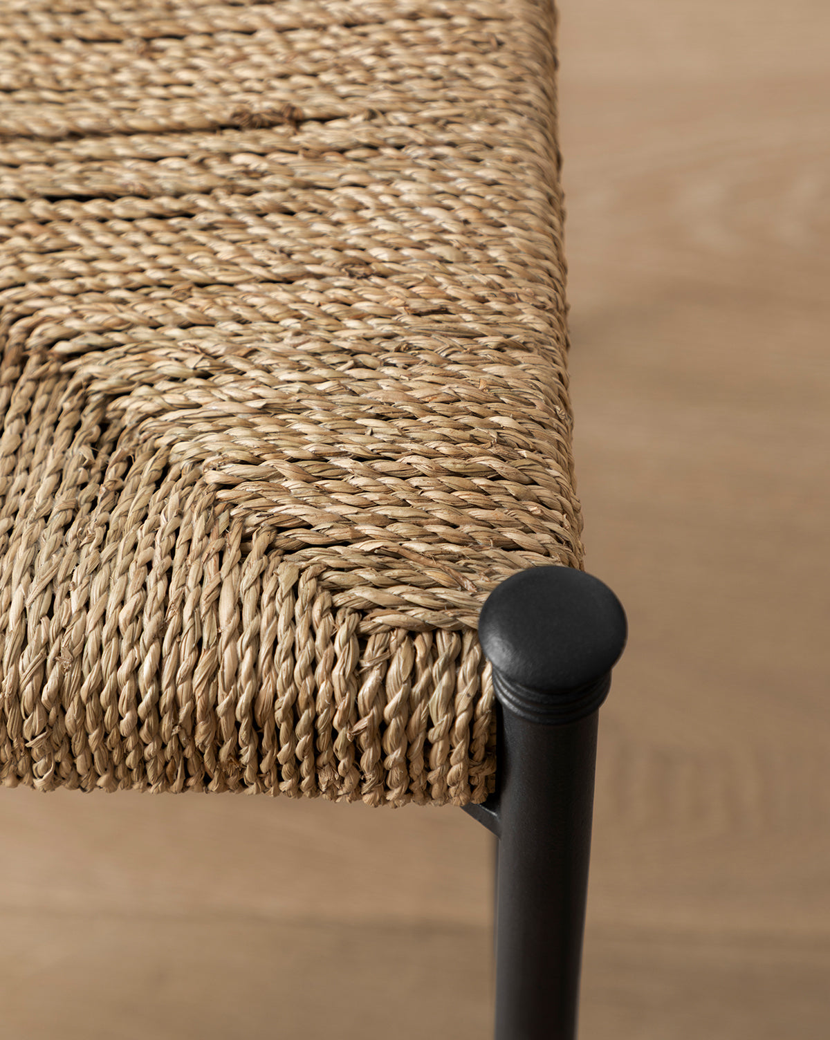 Certainly! Here’s your blended sentence: Close-up of the Makerspalm Peregrine Stool’s woven seagrass seat and black metal legs, set against a smooth, light wood floor, Vendixs