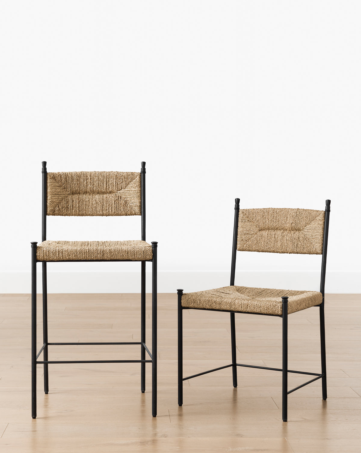 Two Peregrine Stools by Makerspalm, featuring black steel frames and woven straw seats and backs, are displayed on a light wood floor against a plain white background. One stool is taller with a higher seat and backrest than the other. Vendixs