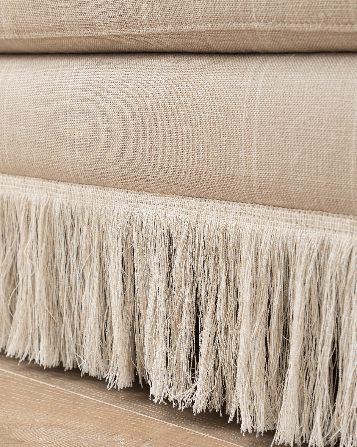 The Philippa Settee (Ready to Ship) features beige woven fabric with visible texture and a white fringe detail at the bottom, adding a stylish touch above a wooden floor, by McGee & Co. The soft, slightly uneven fringe enhances its modern appeal.