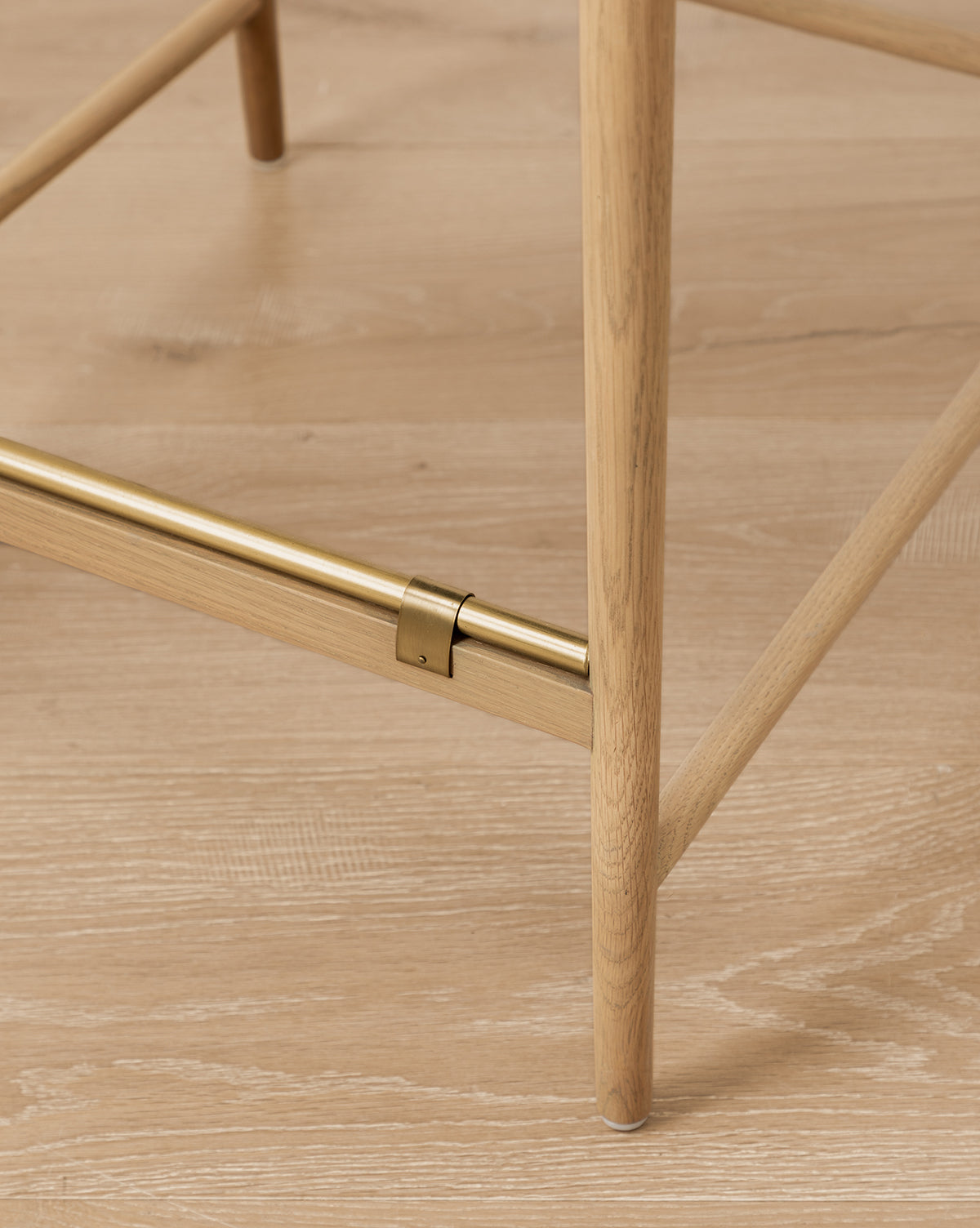 Close-up of a Phipps Woven Bench leg in honey oak with a horizontal brass support bar, standing on a light wood floor. The bench features a woven seagrass seat by McGee & Co.