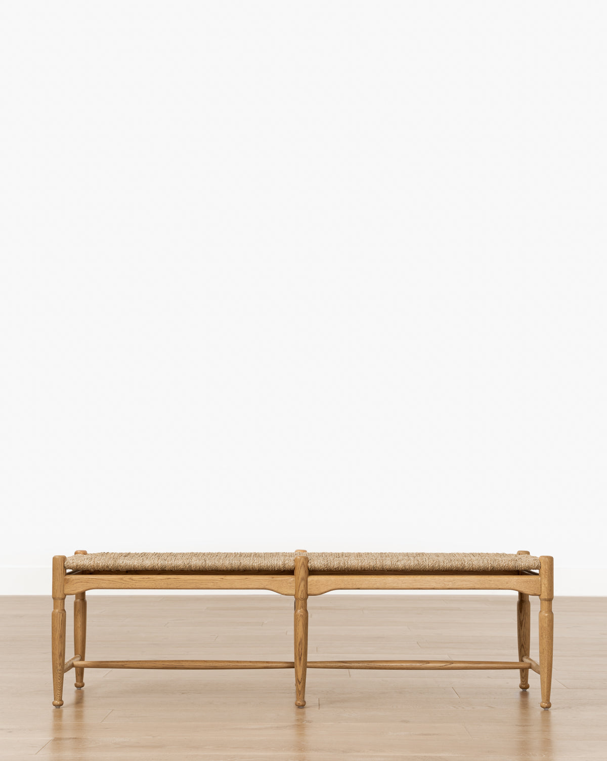 The Phipps Woven Bench, featuring a seagrass seat and wooden frame, is centered on a light honey oak floor against a plain white wall, by Vendixs