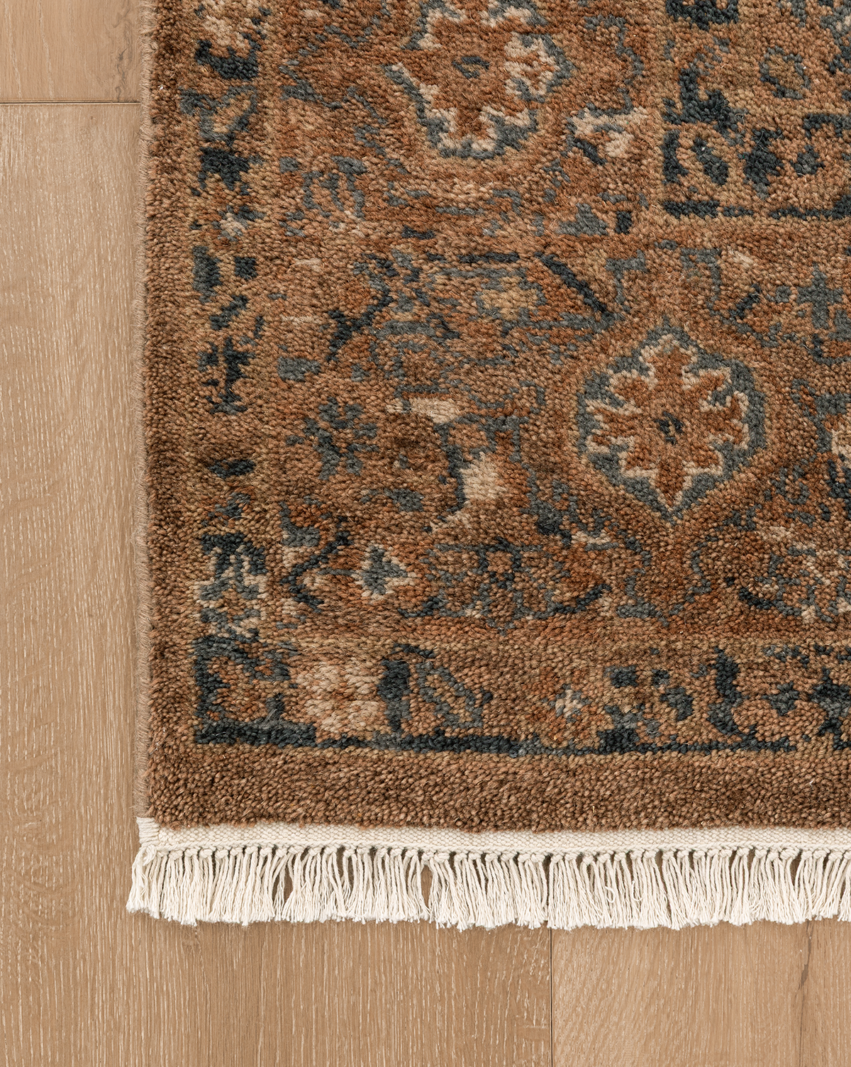 A close-up of the Piedmont Hand-Knotted Wool Rug, showing its brown and blue traditional motifs with white fringes, displayed on a light wooden floor, by Vendixs