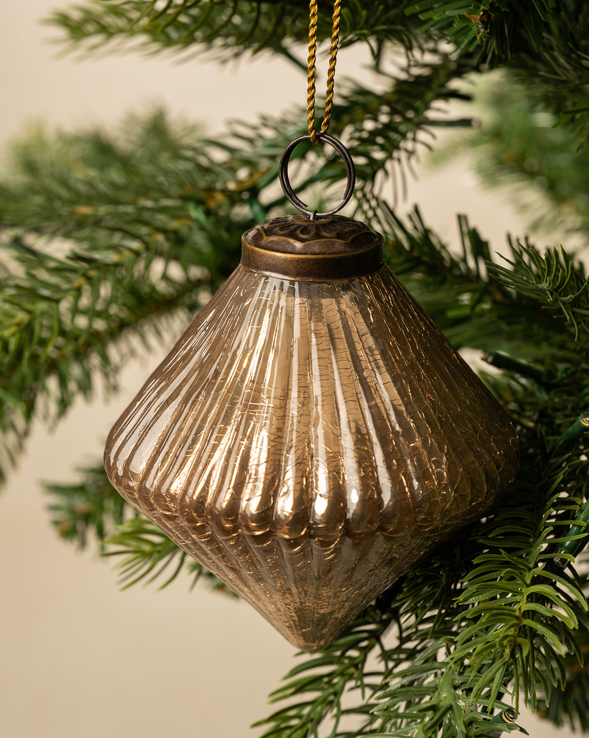 The Piper Ornament, a gold ribbed glass diamond, hangs from a green Christmas tree branch by a gold string, set against a soft neutral background, by Vendixs