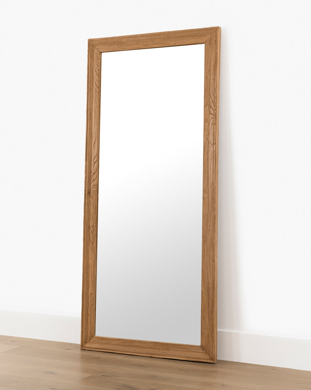 The Pippen Floor Mirror, featuring a large rectangular frame in a honey elm finish, leans against a white wall on a wooden floor, by Vendixs