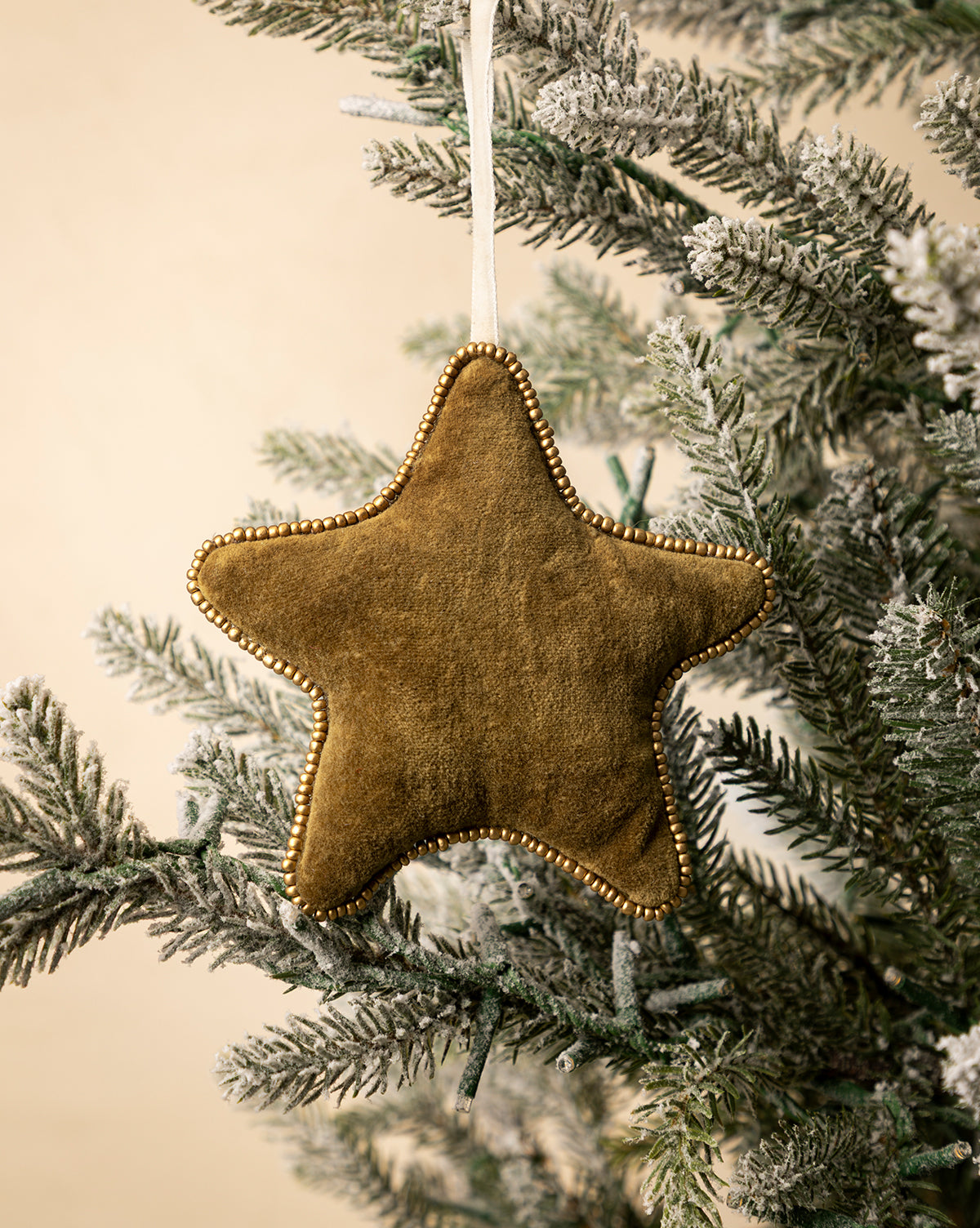 The Plush Beaded Star Ornament, adorned with gold beads and festive design, hangs from a snowy holiday tree branch against a light beige background, by Vendixs
