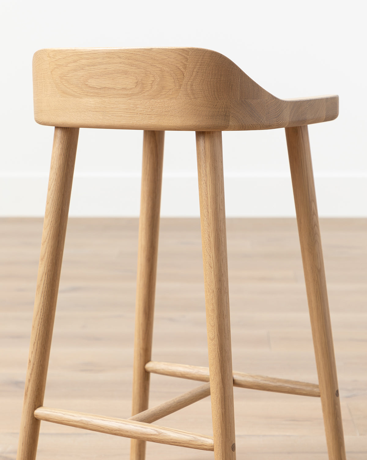 The Makerspalm Portland Counter Stool, featuring four legs and a curved low backrest, sits on a light wood floor against a white wall—perfect for adding style to any kitchen. Vendixs