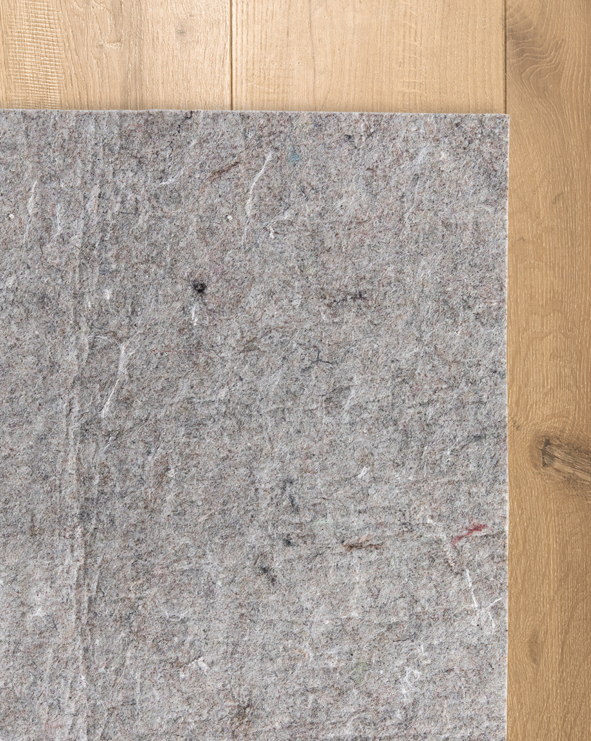 A Premium Indoor Rug Pad lies on a wooden floor, covering part of the surface. Its grey felt features a rough, fibrous texture with visible colored fibers, providing added protection for your floors, by Vendixs