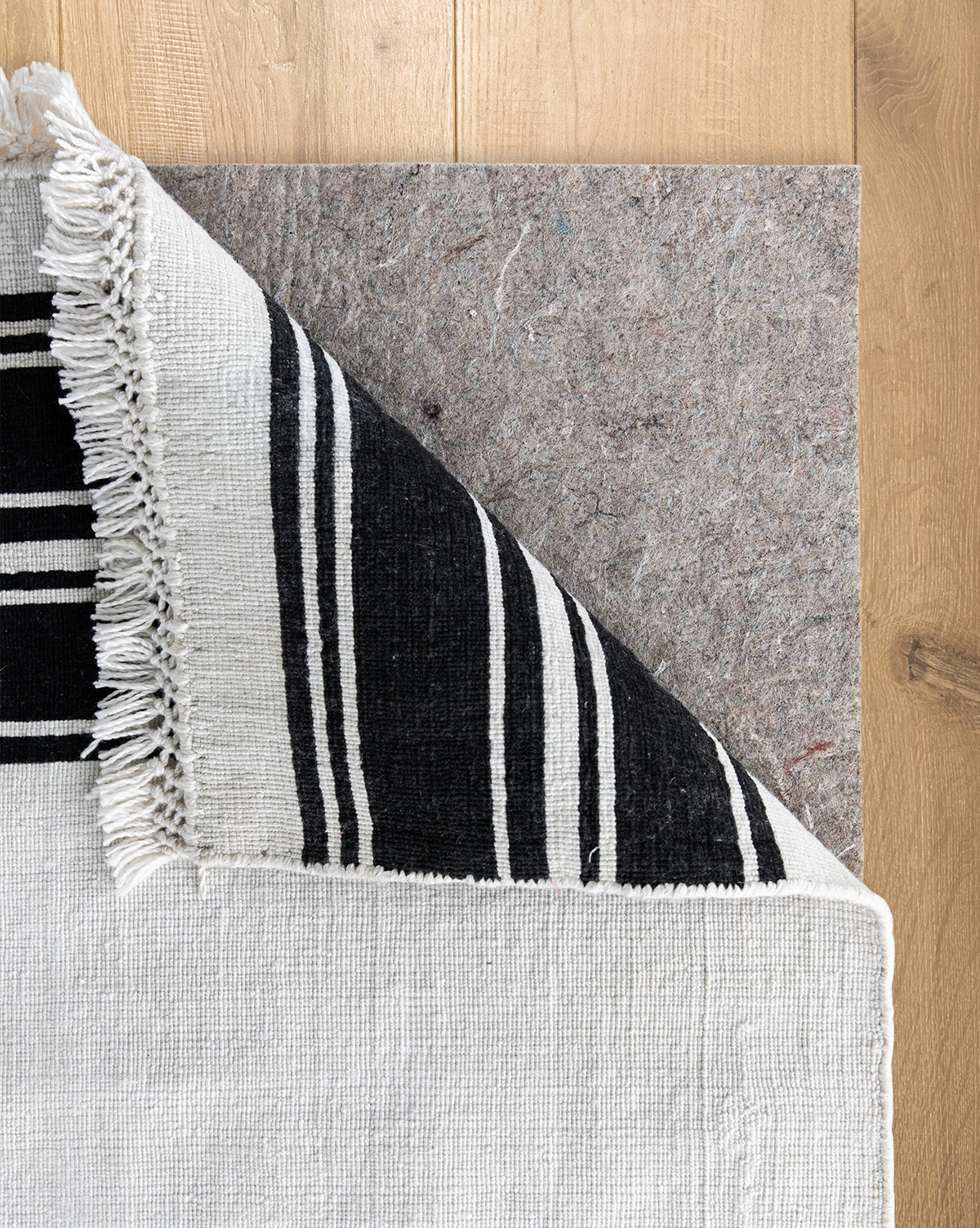 A black and white woven rug with fringed edges is folded at the corner, showing a Premium Indoor Rug Pad with no-slip grip for floor protection on a light wood floor, by Vendixs