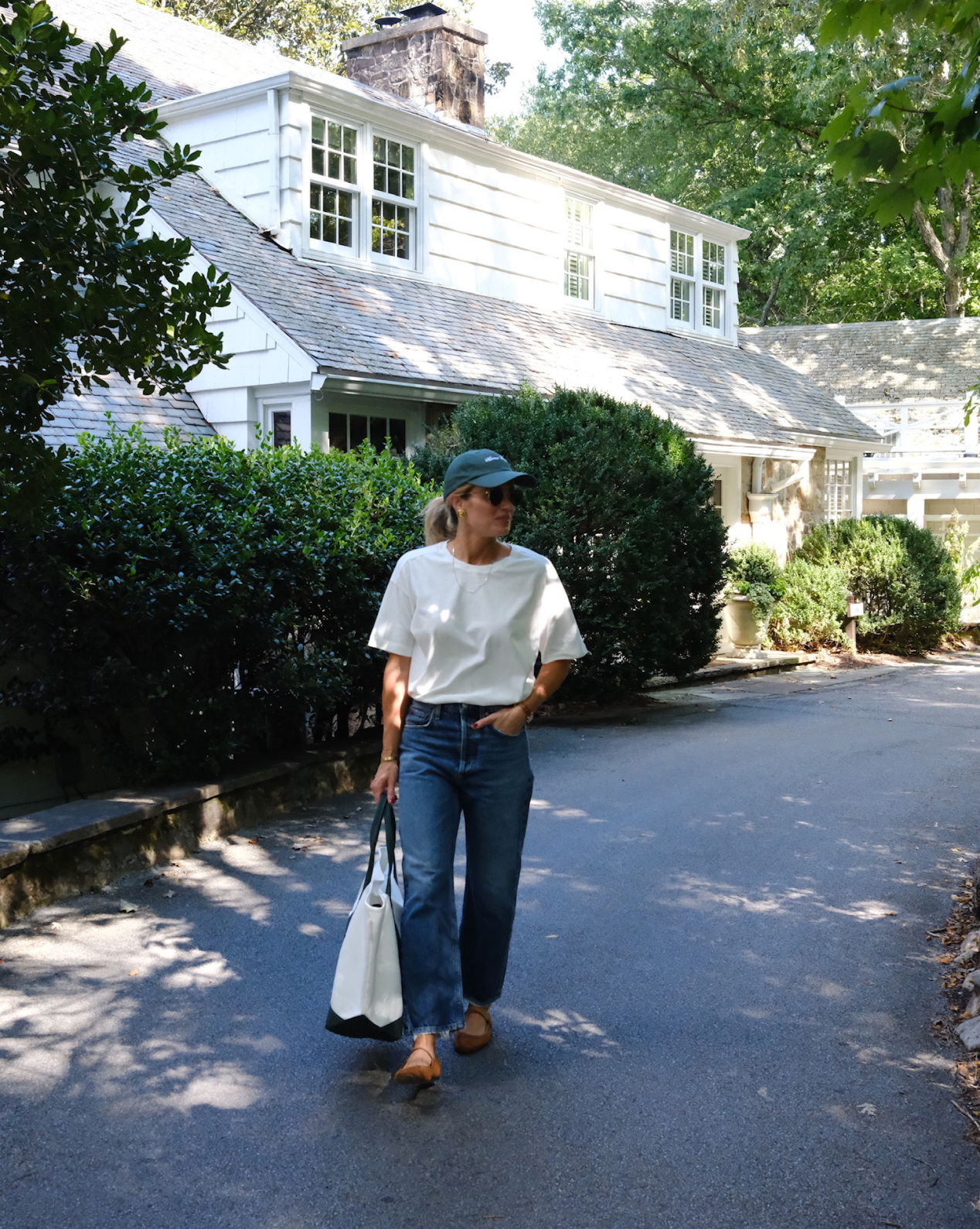 A person in a white t-shirt, blue jeans, green cap, and brown shoes walks on a shaded driveway, carrying the Canvas Laurel Tote from Studio McGee, with a white house and green bushes in the background, by Vendixs