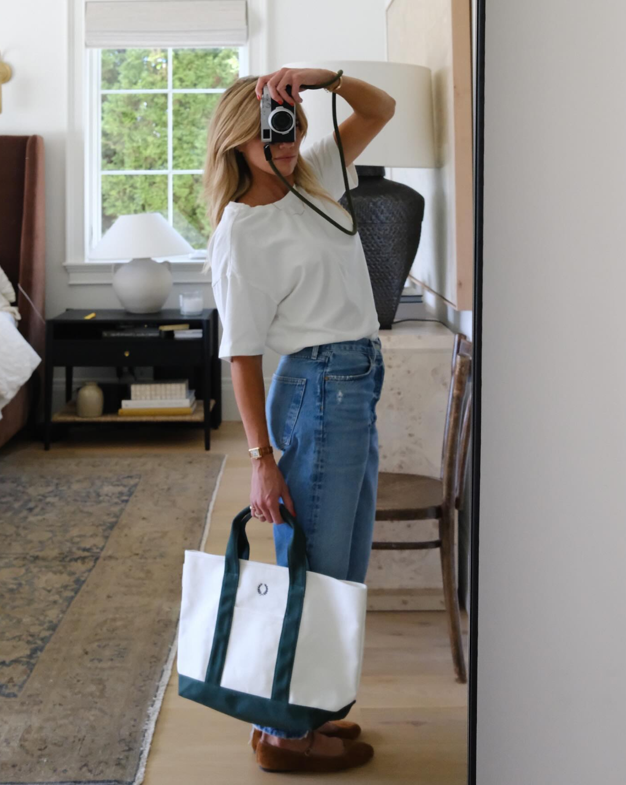 A woman stands in a Studio McGee-inspired bedroom taking a mirror selfie. She wears a white shirt, blue jeans, brown shoes, and carries the Canvas Laurel Tote. The room features a bed, rug, chair, and window, by Vendixs