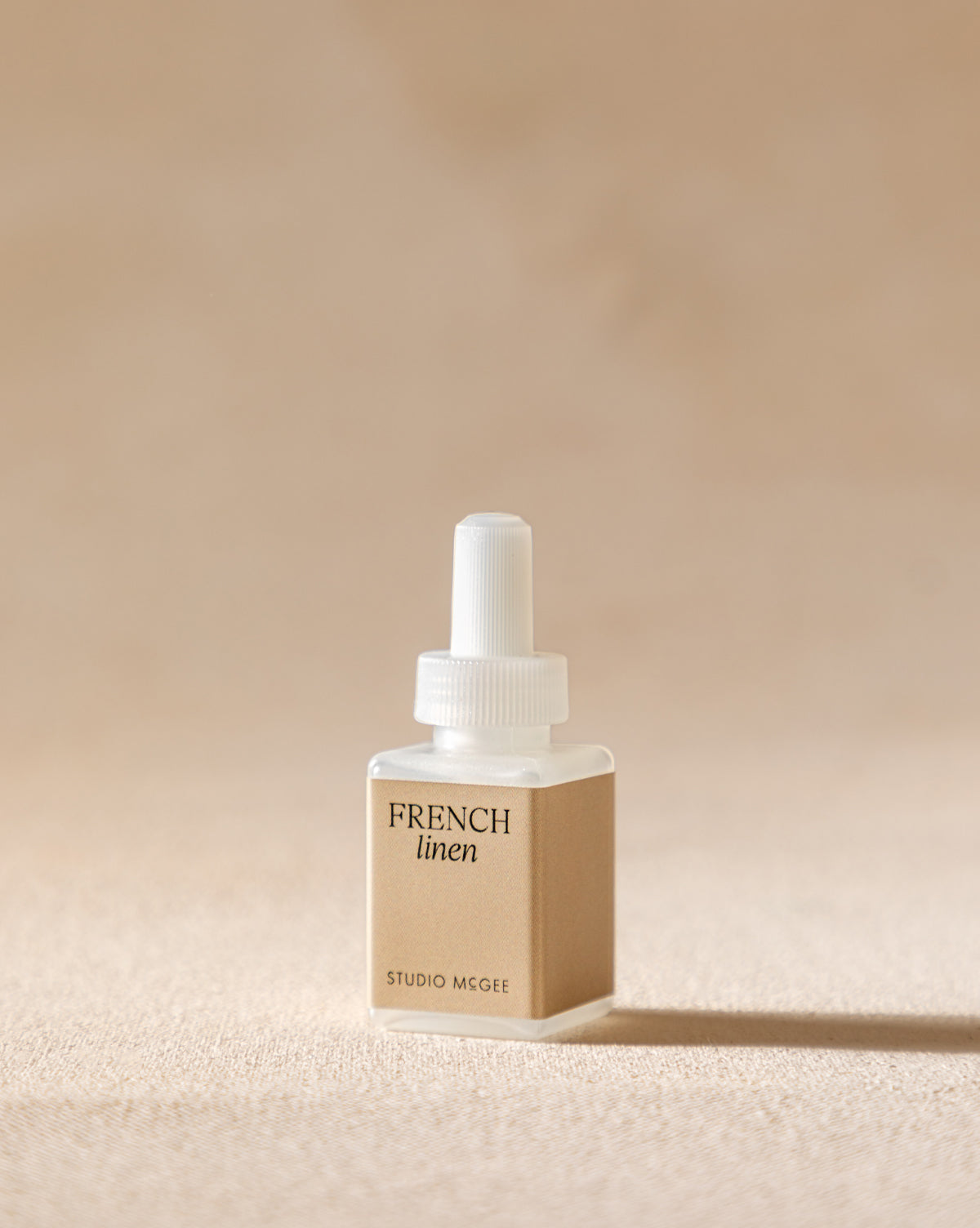 A small, square Pura x Studio McGee French Linen Home Fragrance Refill stands upright on a beige surface, featuring a white cap and dropper against a softly blurred neutral background, suggesting its fresh citrus scent.
