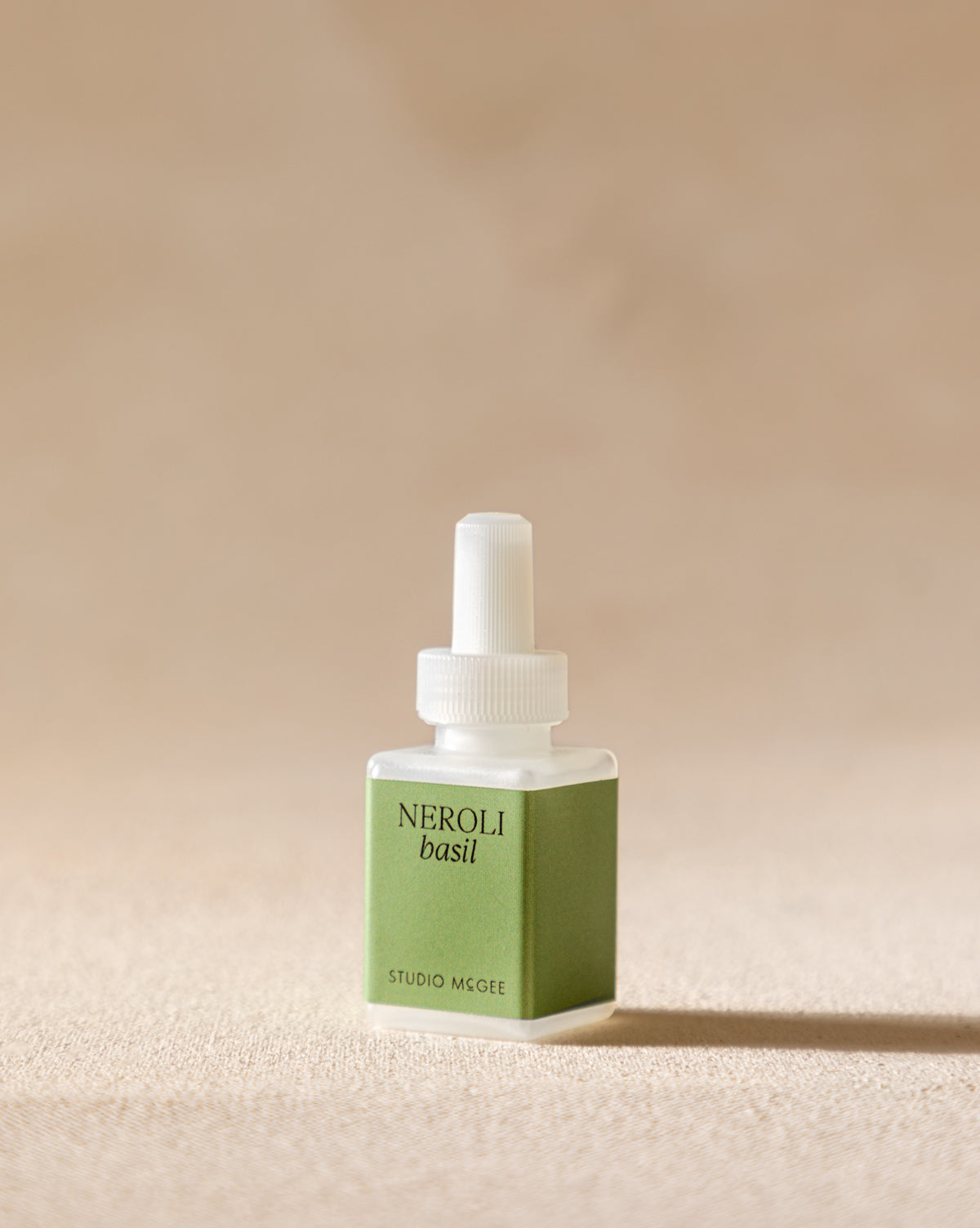 A small square bottle of Pura x Studio McGee Neroli Basil Home Fragrance Refill, with a green label and white cap, rests on a beige surface, evoking an aromatic blend of herbal and citrus notes.