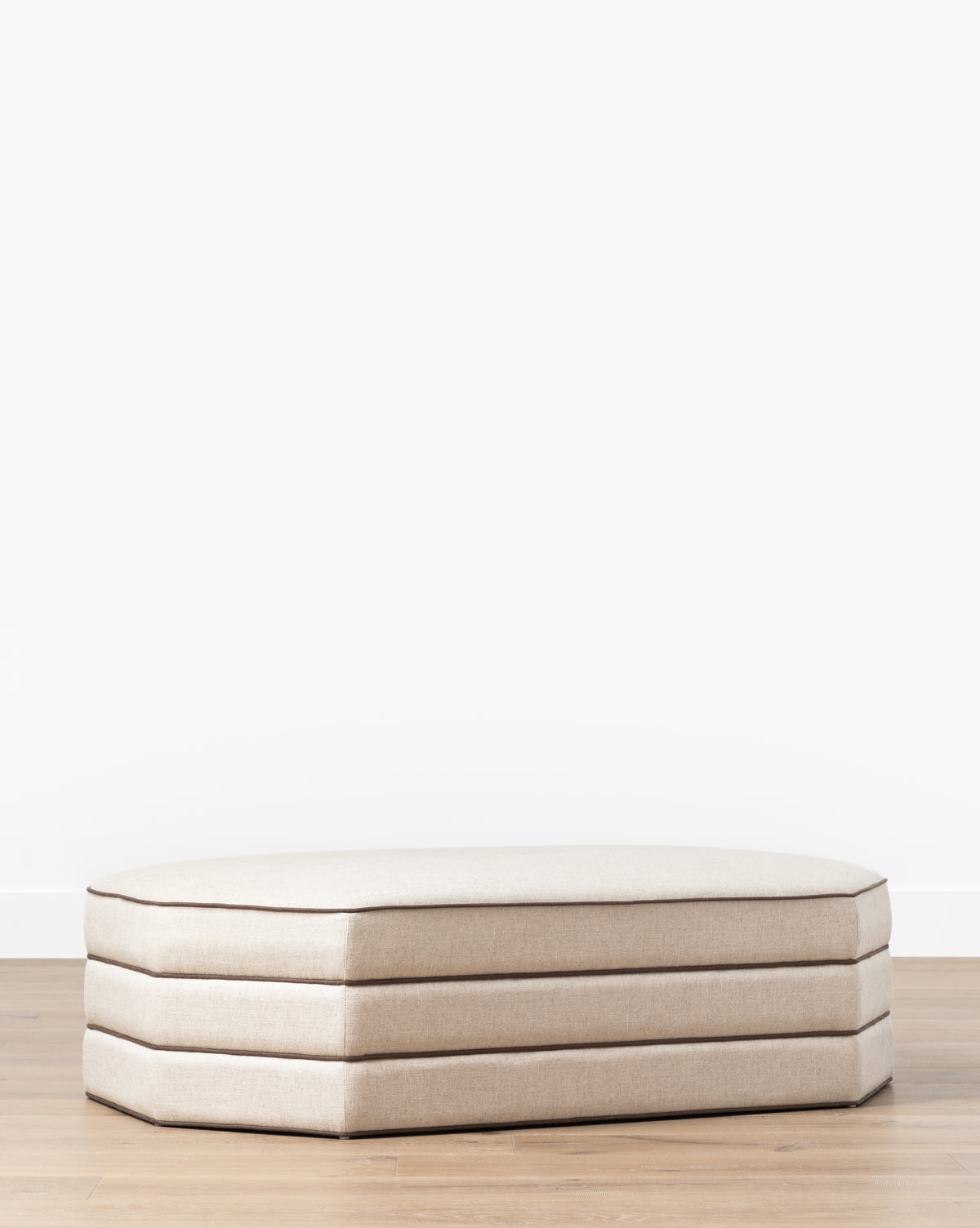 A Makerspalm Rafferty Ottoman in beige performance linen with three stacked cushions rests on a light wood floor against a plain white wall, Vendixs