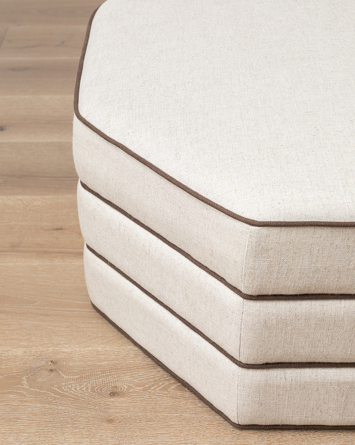 A stack of three beige Rafferty Ottomans by Makerspalm, hexagonal with brown piping and performance linen upholstery, arranged on a wooden floor, Vendixs