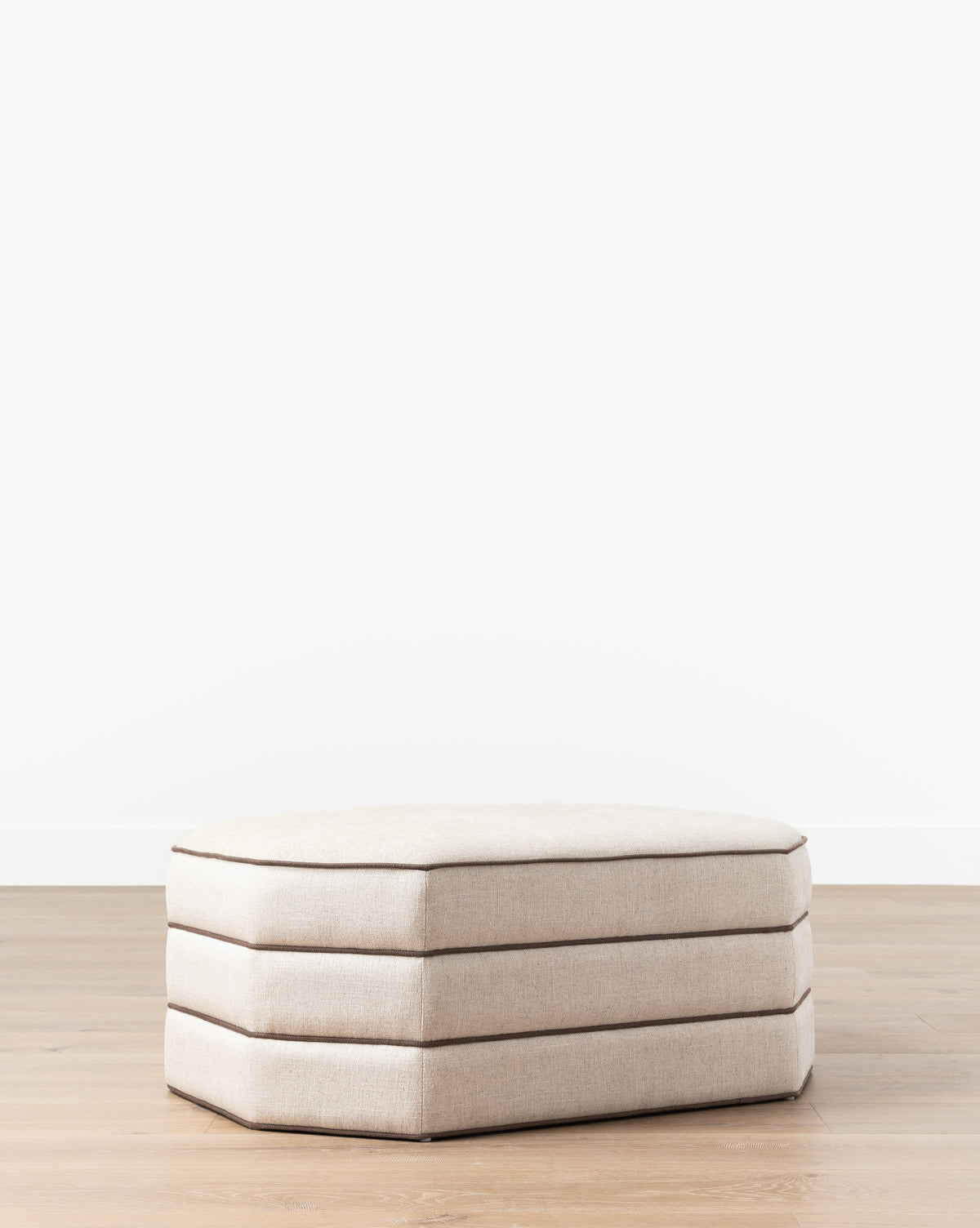 The Makerspalm Rafferty Ottoman, upholstered in beige performance linen with three stacked cushion layers, stands on a light wooden floor against a plain white wall. Vendixs