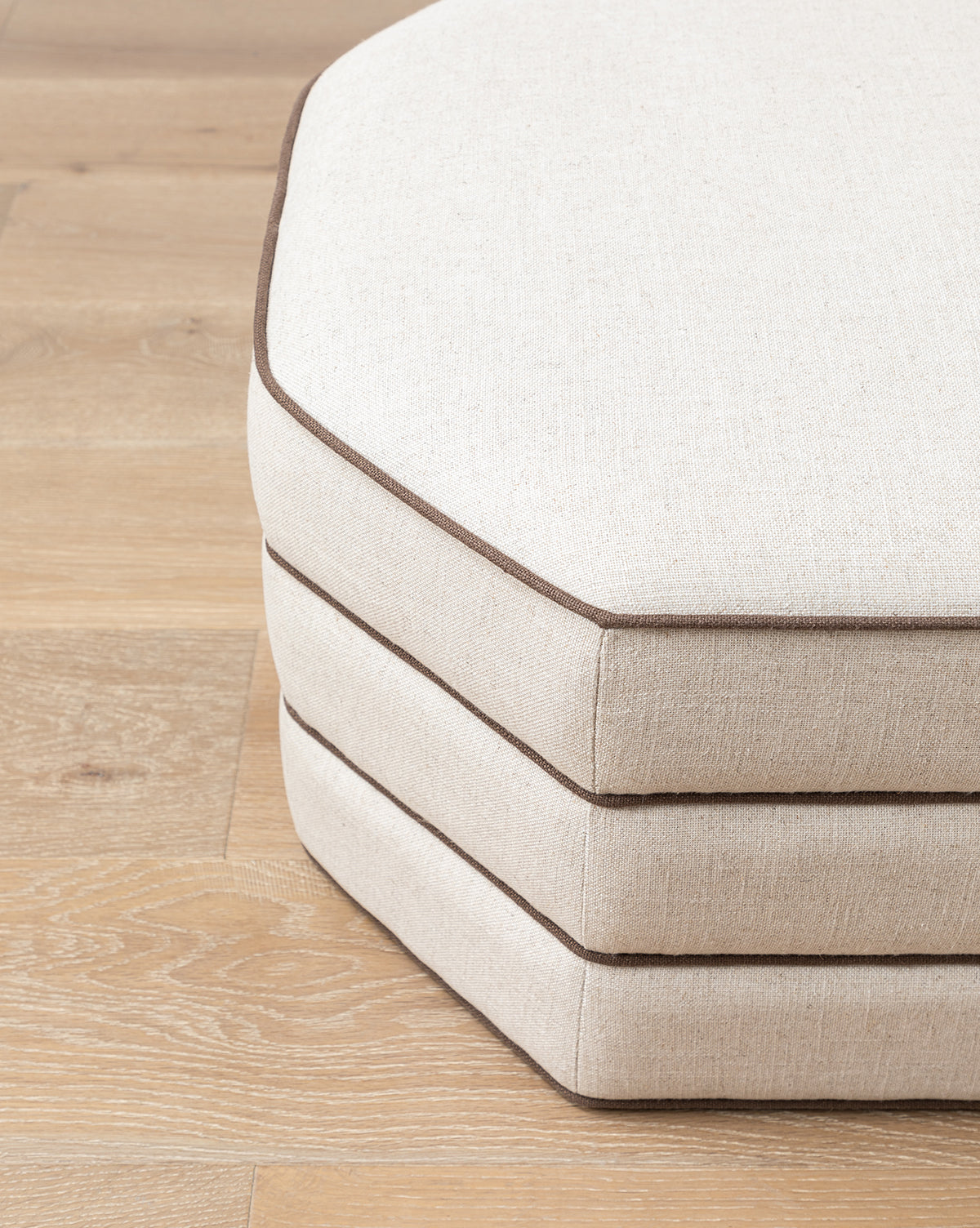 Three beige, octagonal Makerspalm Rafferty Ottoman cushions with dark brown piping in performance linen are stacked on a light wooden floor, Vendixs