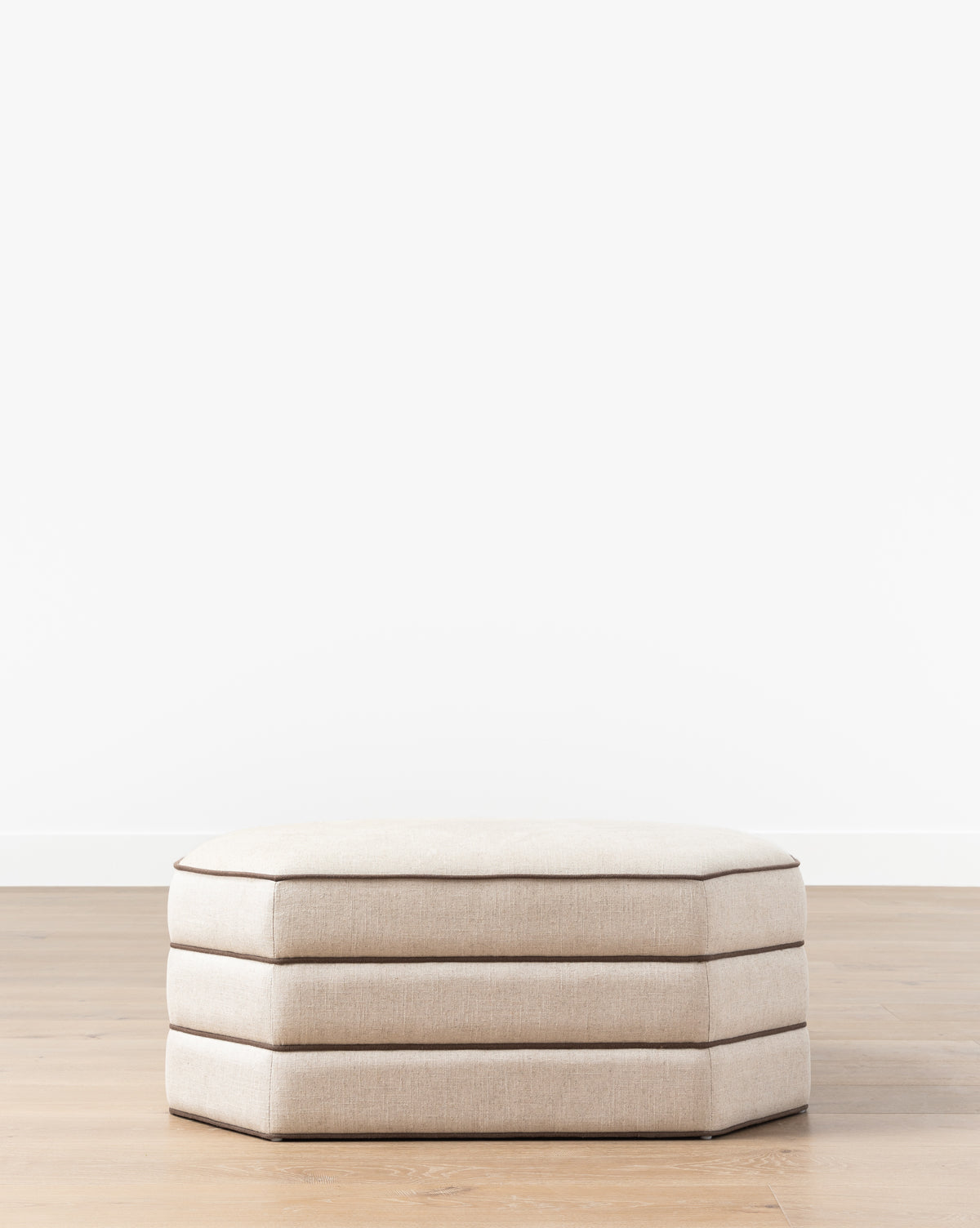 The Makerspalm Rafferty Ottoman, a beige rectangular piece with layered performance linen cushions, is set on a light wood floor against a plain white wall. Vendixs