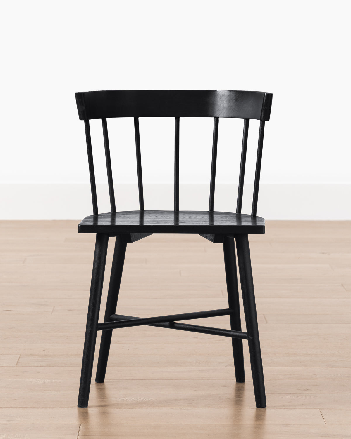 The Reeves Dining Chair, featuring a sleek black finish, curved backrest, and vertical slats, stands on a light brown wooden floor against a plain white wall, by Vendixs