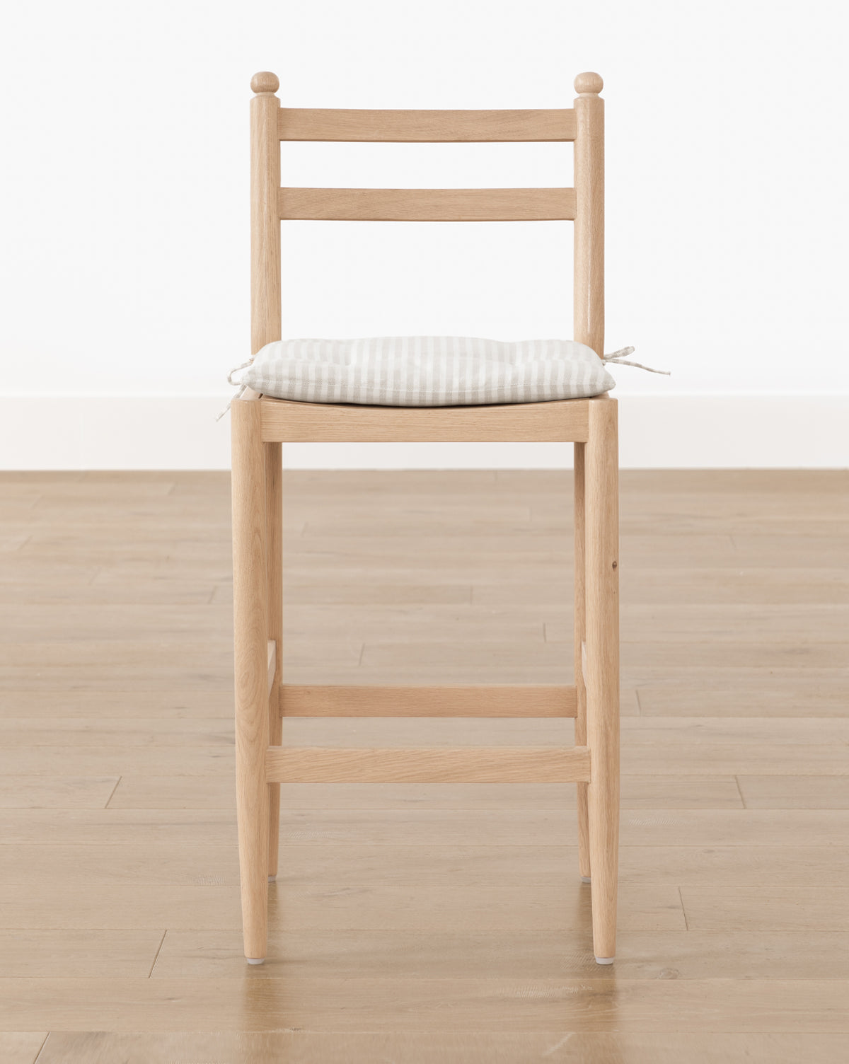 The Romany Counter Stool, with straight wooden legs and a ladder backrest, features a light checkered seat cushion tied to the seat and sits on a light wood floor against a plain white wall, showcasing modern simplicity and style, by McGee & Co.