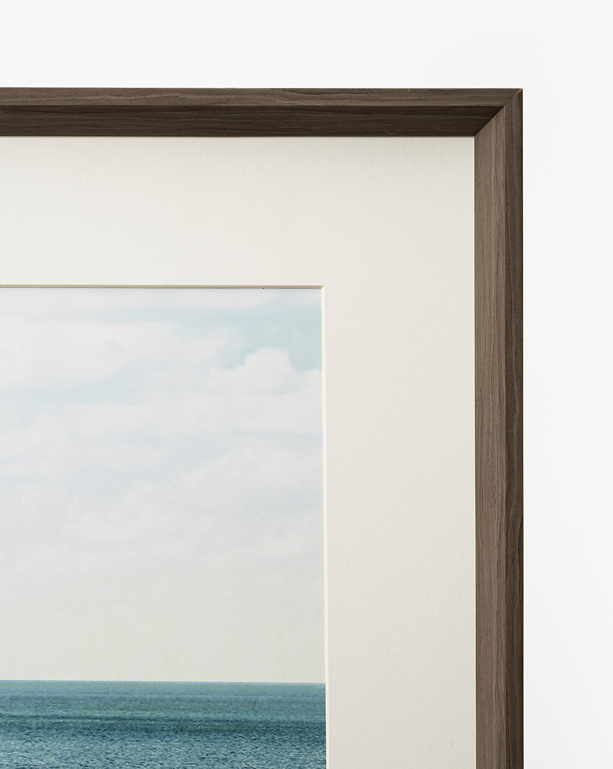 A close-up of the top right corner of Room with a View, a gallery-quality art piece showing a cloudy sky, blue ocean, off-white mat, and dark walnut frame set against a white background by Vendixs