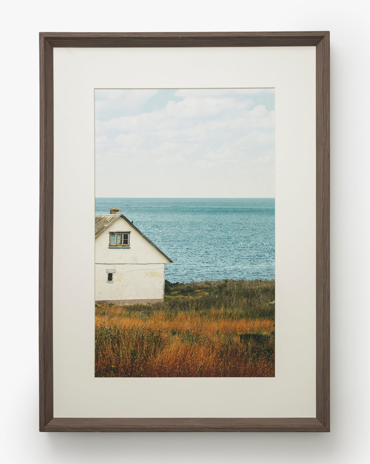 Room with a View: A gallery-quality framed photograph of a white house by the sea, tall grasses in front, and a calm blue ocean under cloudy skies, all set in a dark walnut frame, by Vendixs