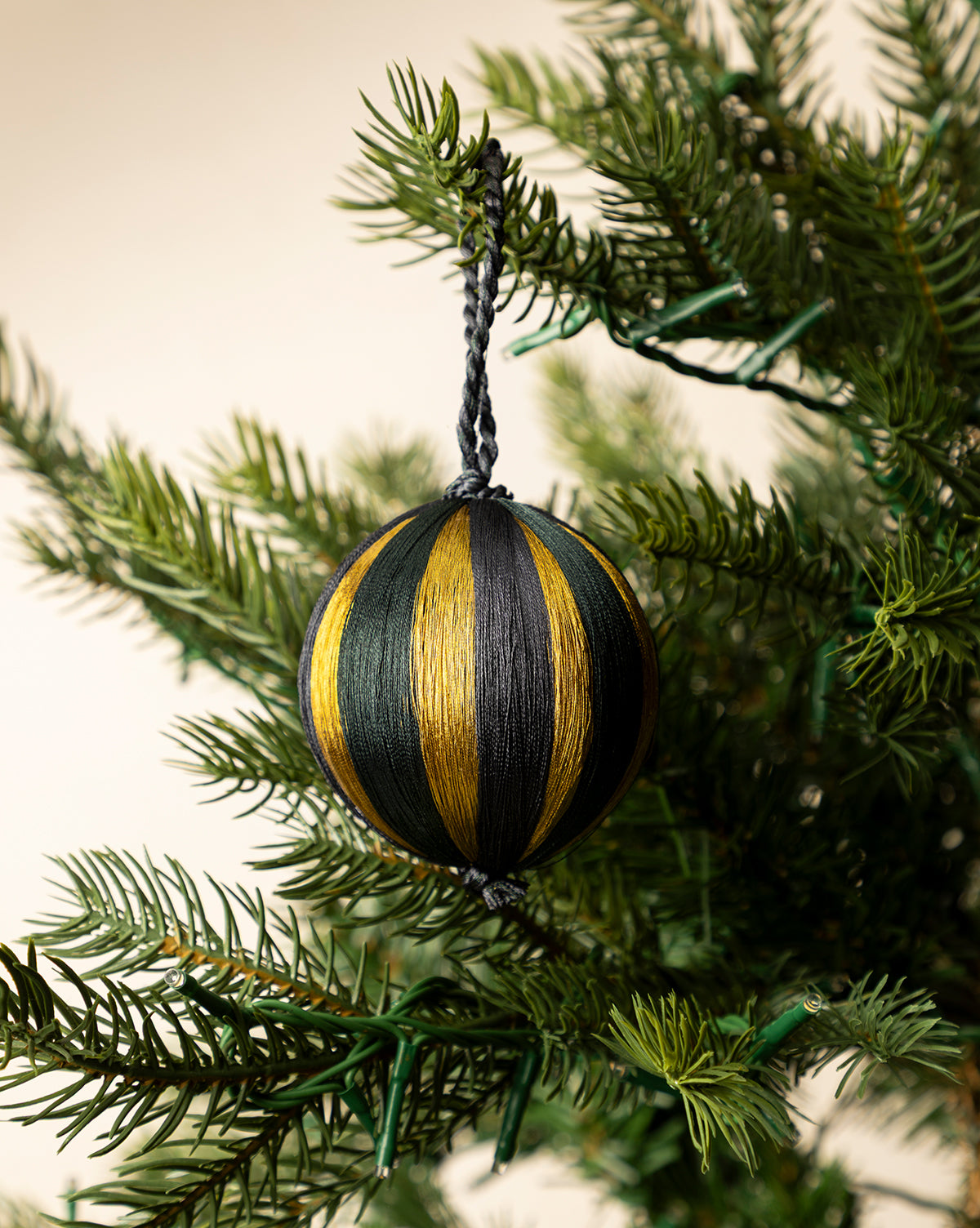 A 3" Satin Threaded Bauble in black and gold stripes hangs from a green Christmas tree branch, with holiday lights peeking through the needles, by Vendixs
