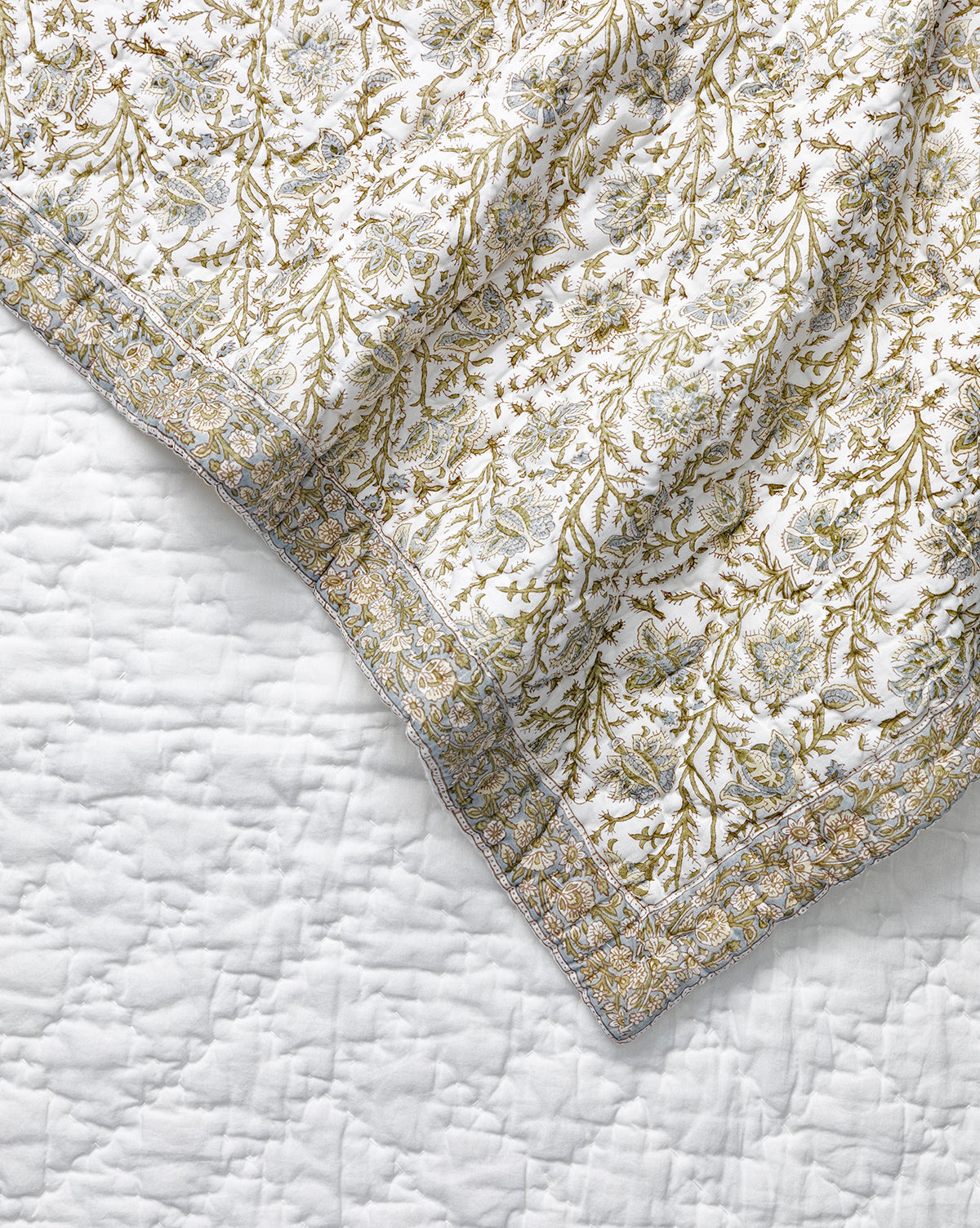 A close-up of the Seton Kids Quilt by Renuka Inc., featuring a leafy floral pattern in green and gold on white, draped over a plain white textured bedspread, Vendixs