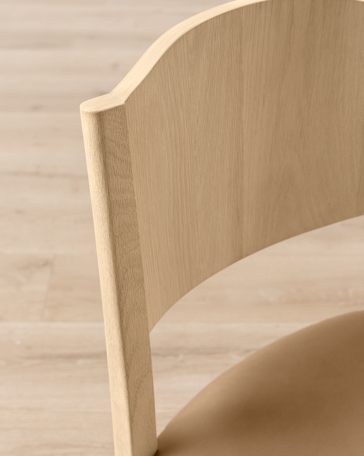 Close-up of the Shepherd Counter Stool by Makerspalm, showing its smooth, curved white oak backrest and seat with natural wood grain, set on a matching wood floor. Vendixs