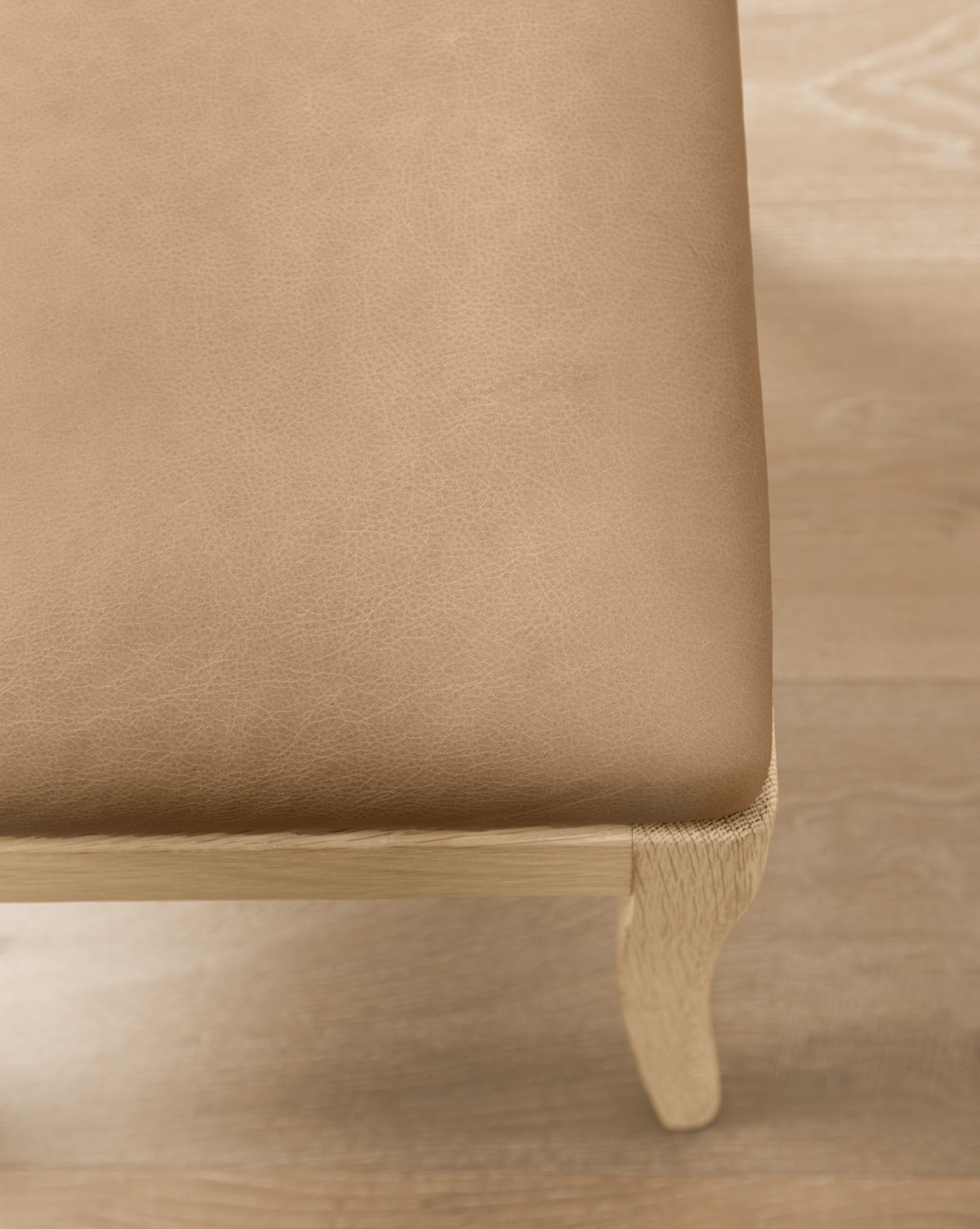 Close-up of the Shepherd Counter Stool by Makerspalm, featuring a light brown leather seat cushion with smooth performance upholstery and a curved white oak leg, all set on a light wood floor. Vendixs
