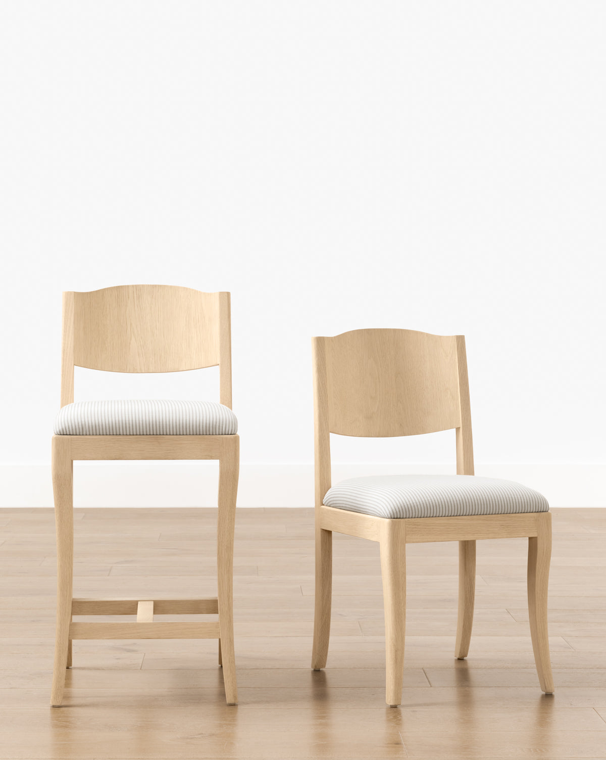 Two light wood chairs, including the Makerspalm Shepherd Counter Stool with a curved back and white-and-gray striped performance upholstery, sit side by side on a light wood floor against a crisp white wall. Vendixs